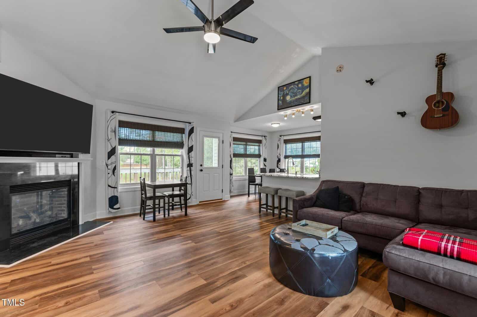 Modern living room with wood flooring, sectional sofa, round coffee table, wall-mounted TV above a fireplace, dining table by windows, and a guitar hung on the wall. Ceiling fan and natural light enhance the space.