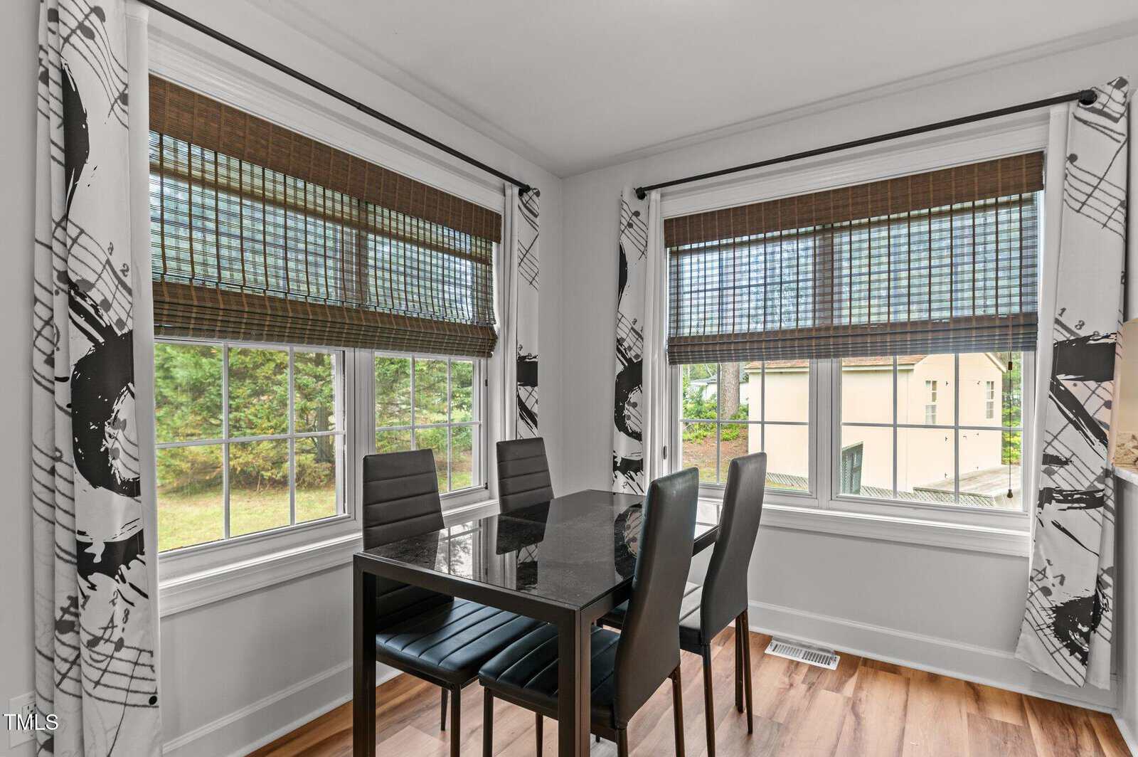 A dining area with a black glass table and four black chairs is positioned by two windows with bamboo shades and patterned curtains. The floor is wood, and the windows offer a view of trees and a building outside.
