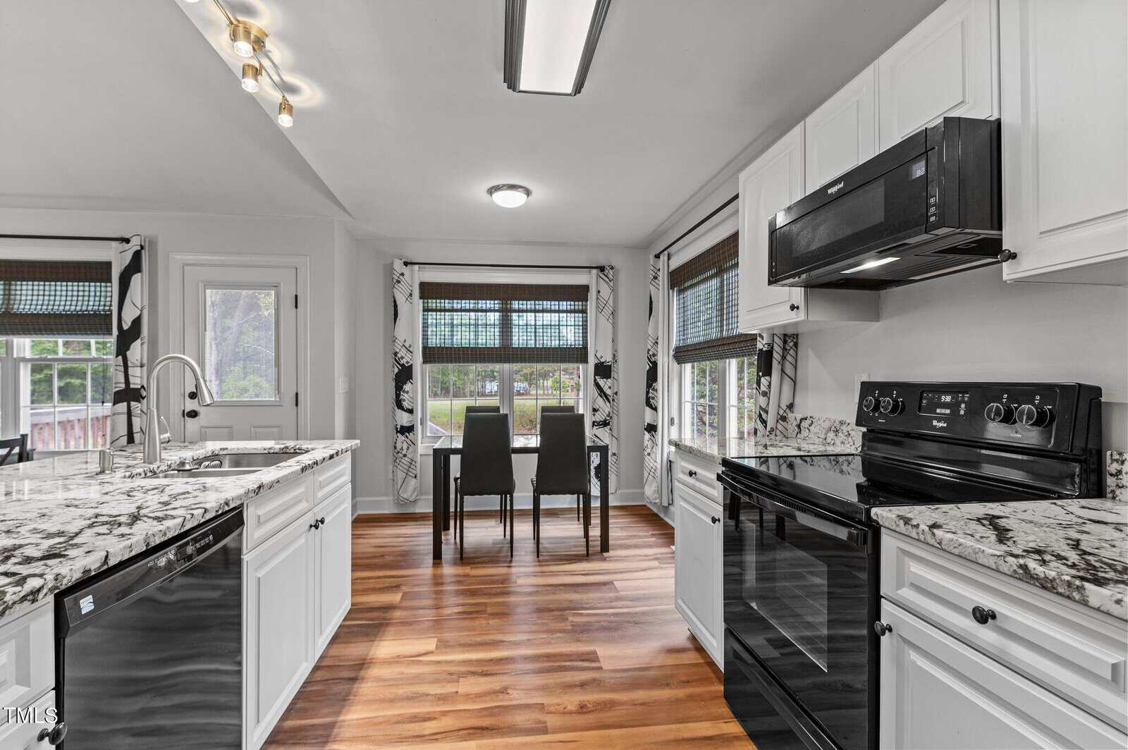 A modern kitchen with white cabinets, black appliances, marble countertops, and wood flooring. A small dining area with a table and four chairs is by windows with patterned curtains, letting in natural light.