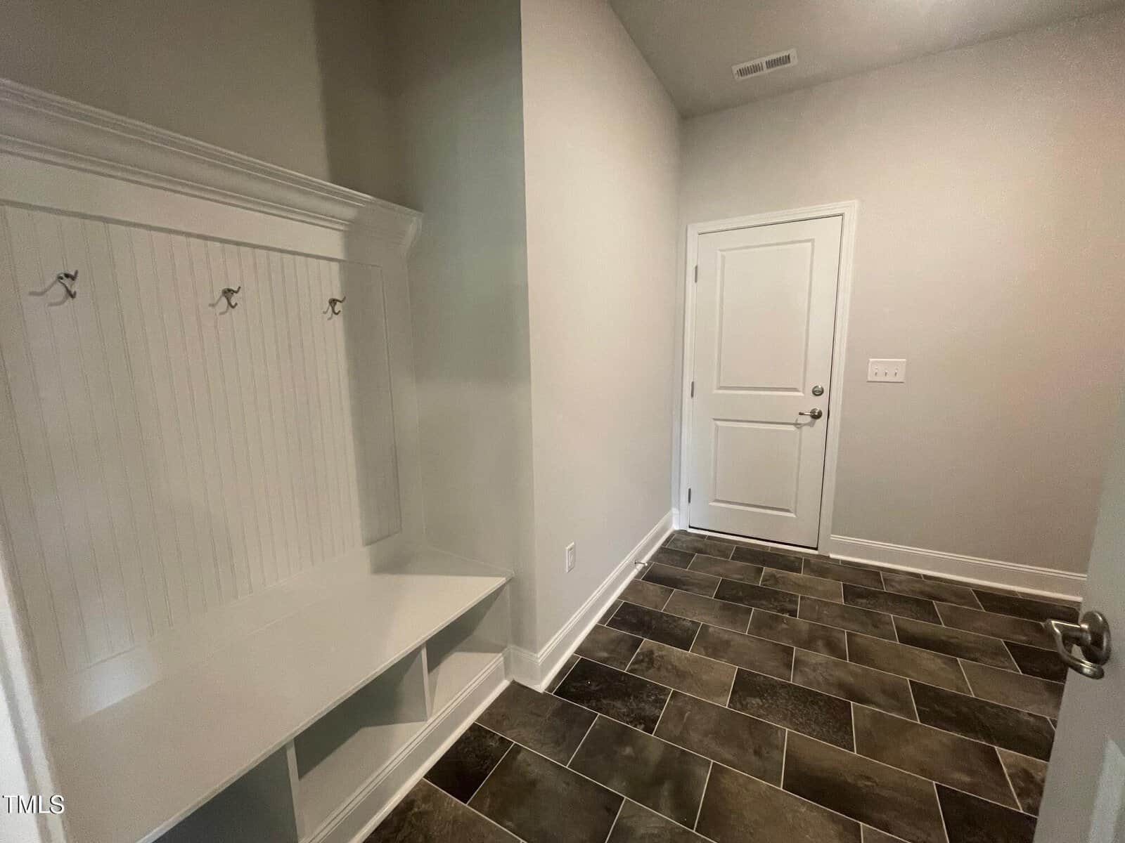 A mudroom with a white bench, built-in coat hooks, and cubbies on the left. The floor has dark, rectangular tiles. A white door and a light switch are on the right wall. The walls are painted light gray.