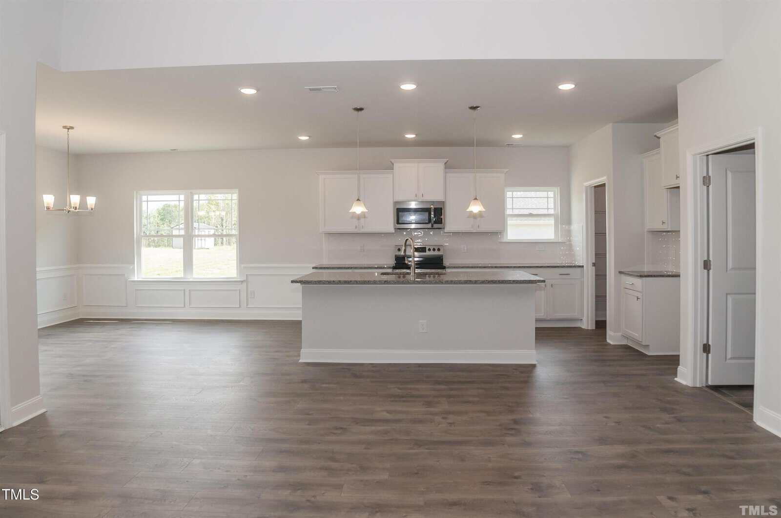 Open concept kitchen and dining area with white cabinets, stainless steel appliances, pendant lights over a large island, and wood-style flooring. Large windows let in natural light. Walls are painted light gray.