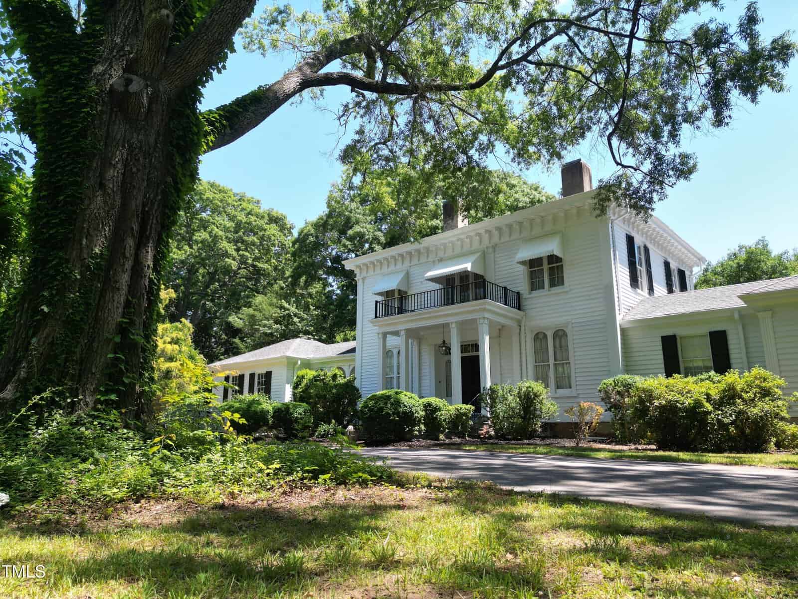 A large white two-story house with columns and black shutters stands among trees and greenery at 508 Eaton Avenue, Warrenton, with a driveway curving in front and sunlight filtering through the branches.