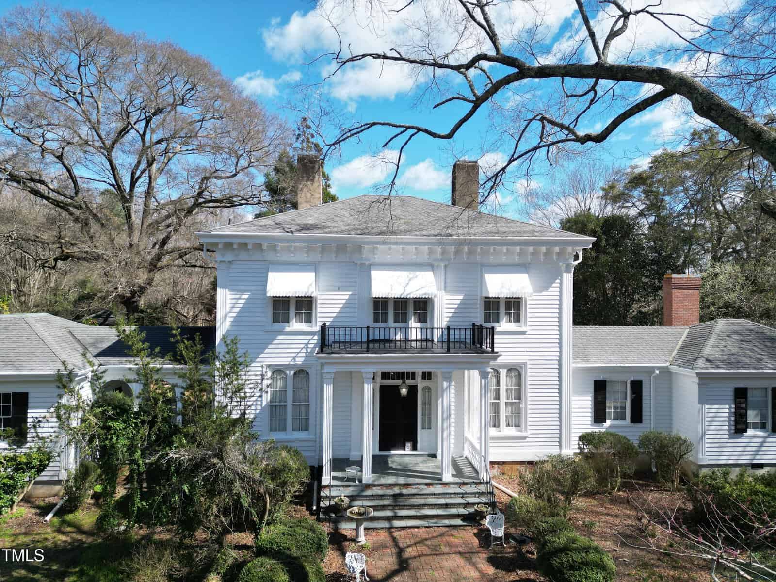 A large white two-story house with a columned front porch, black balcony, and brick walkway, surrounded by trees and landscaping under a blue sky with clouds.