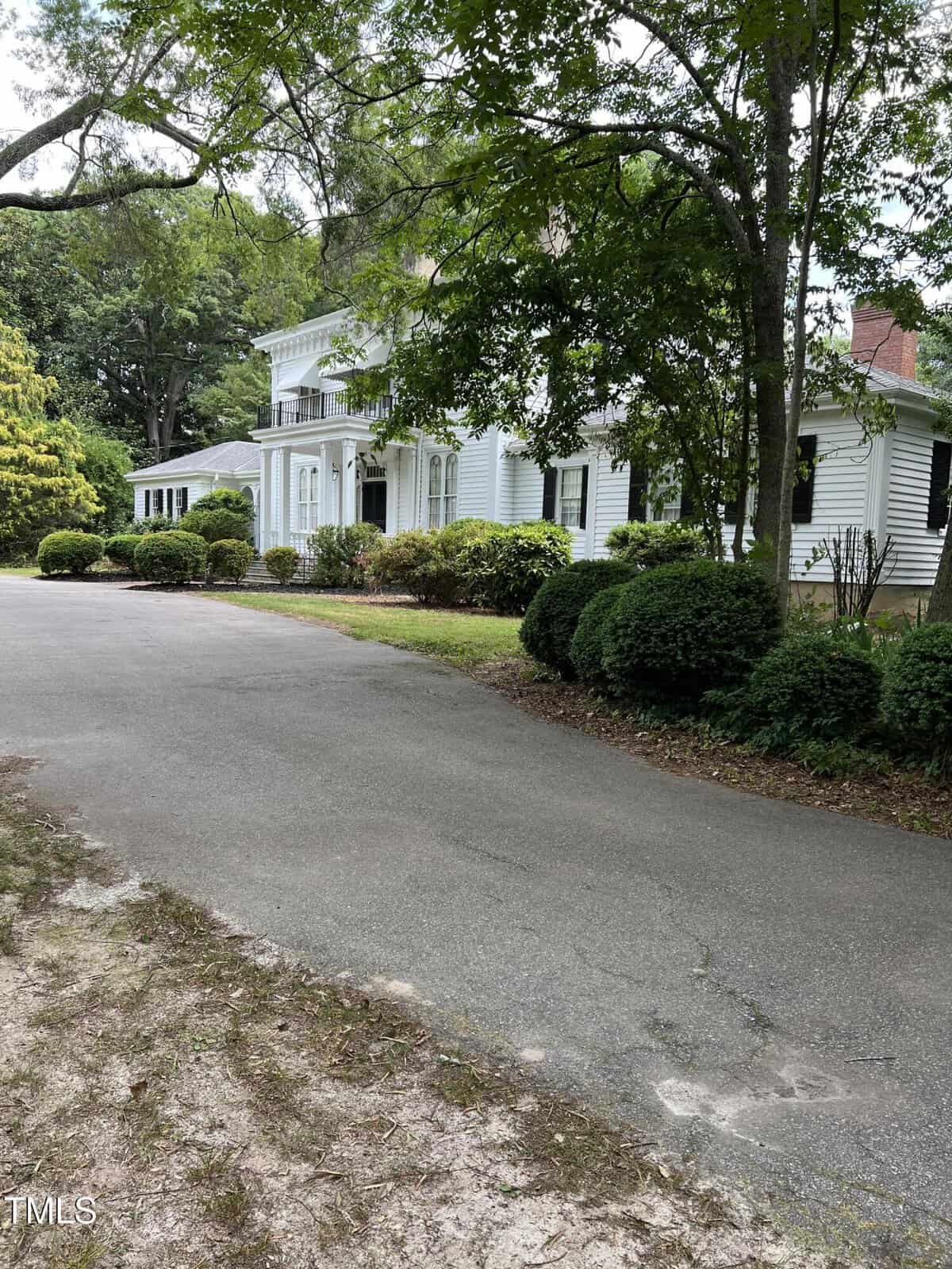 White, two-story house with large pillars and a porch, surrounded by bushes and trees. A paved driveway curves in front, with grass and dirt along the edge. The scene is shaded by overhanging branches.
