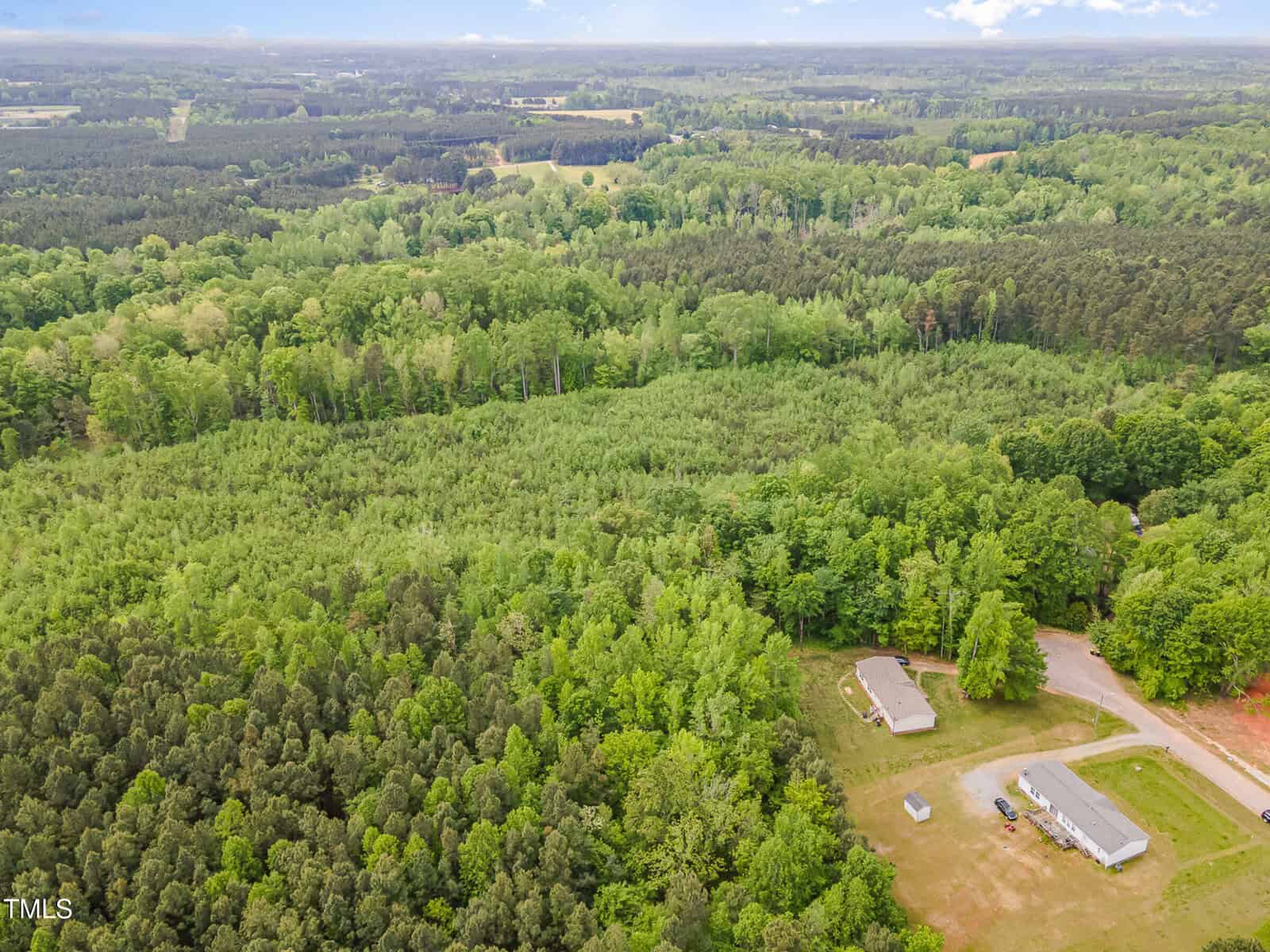Aerial view of a large, densely wooded area with two single-story buildings and a cleared grassy area near a road in the bottom right corner; forest extends toward the horizon.