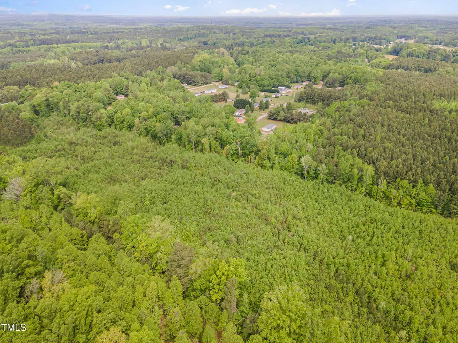 Aerial view of a densely wooded area with a mixture of forests and clearings; several buildings are visible in the midground, surrounded by trees, and a distant horizon under a partly cloudy sky.