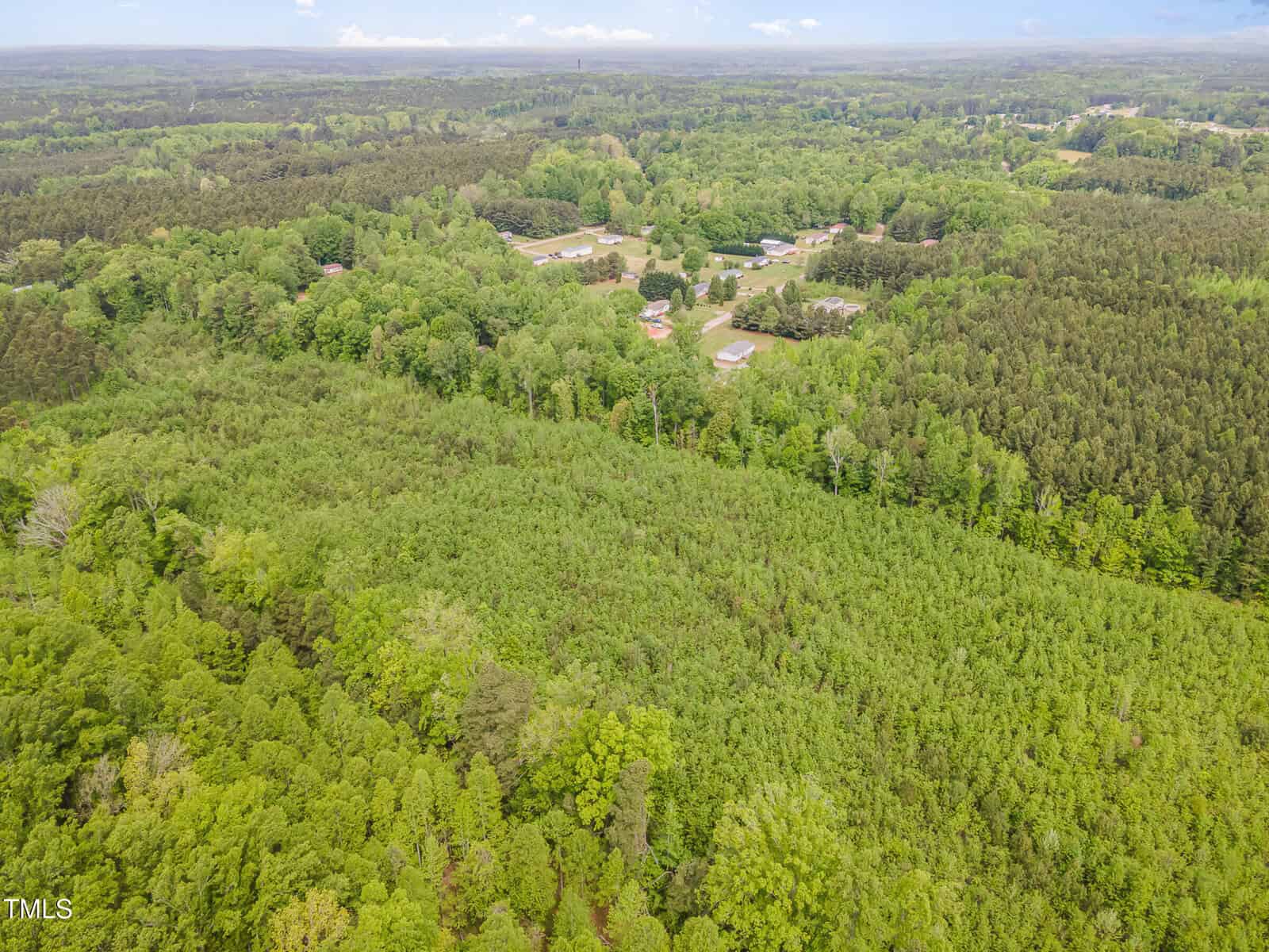 Aerial view of a densely forested area with green trees, with several buildings and open spaces visible in the distance surrounded by more trees. The landscape extends to the horizon under a partly cloudy sky.