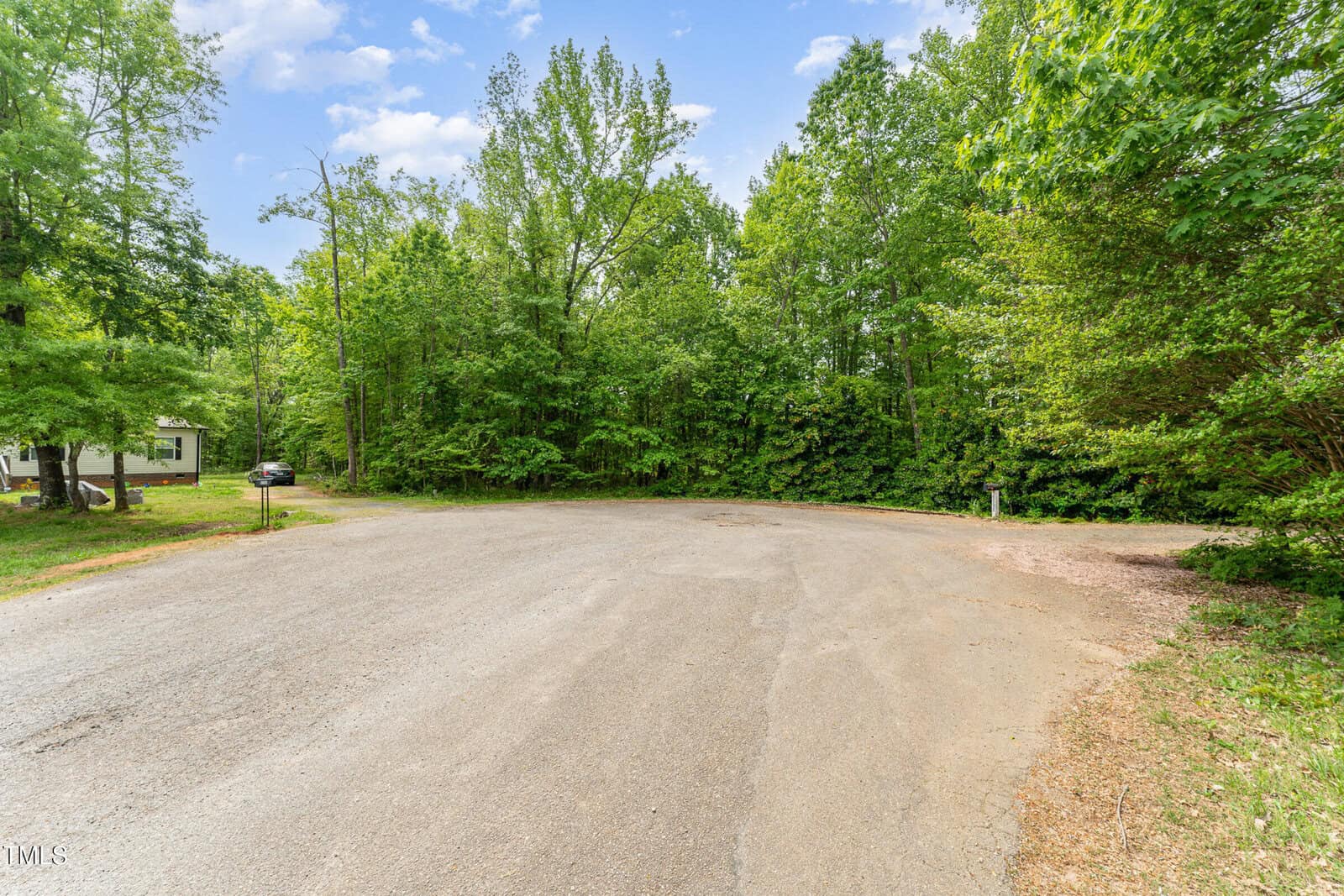 A paved cul-de-sac surrounded by dense green trees, with a mailbox on the right and a parked car partially visible on the left near a house. The sky is partly cloudy.