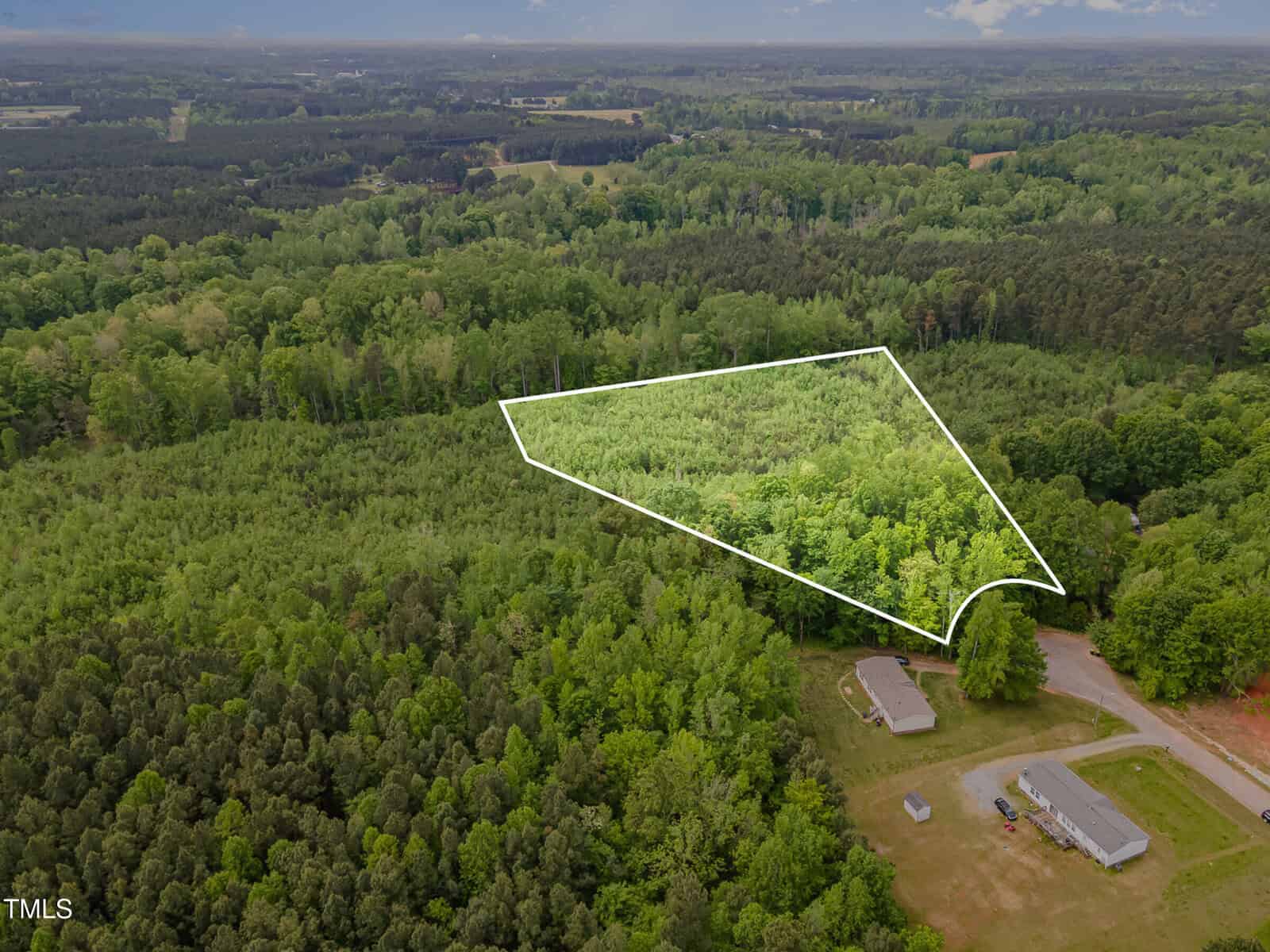 Aerial view of a forested area with a plot of land outlined in white; two buildings and a road are visible in the lower right corner, surrounded by dense trees.