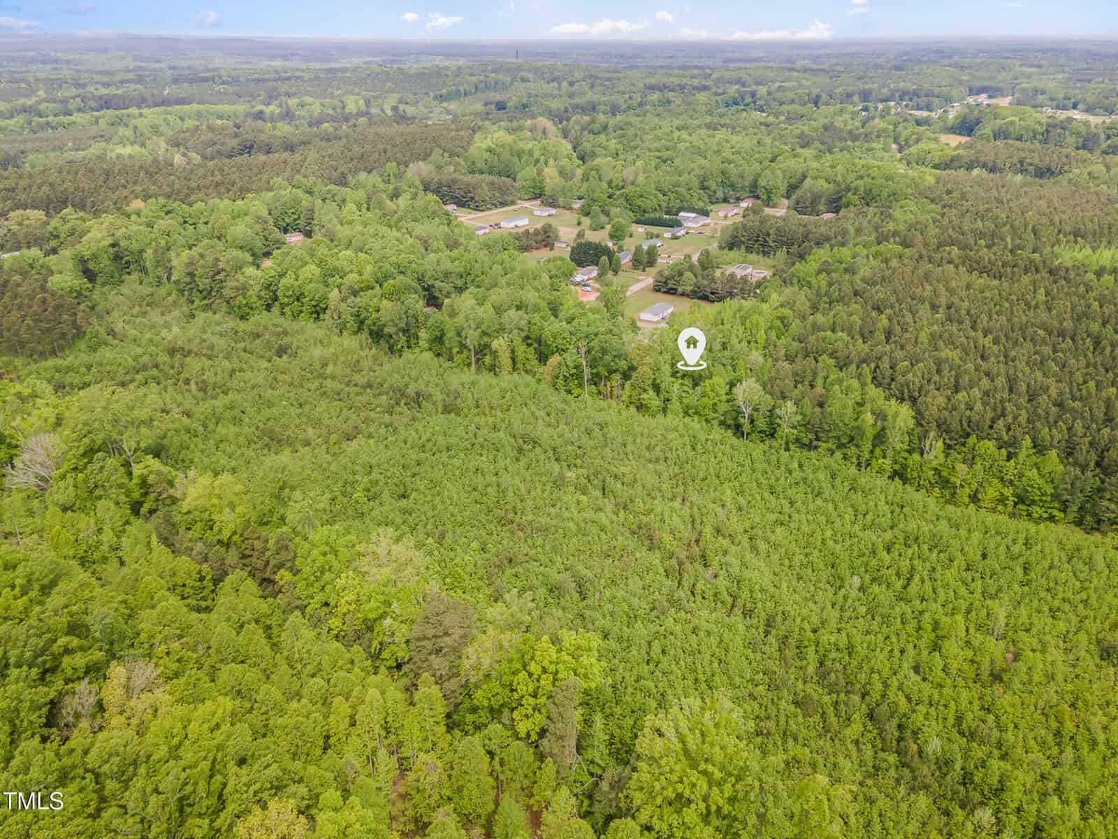 Aerial view of a densely wooded area with several houses and buildings visible in the background. A location marker highlights a specific spot among the trees near the buildings. The landscape is lush and green.