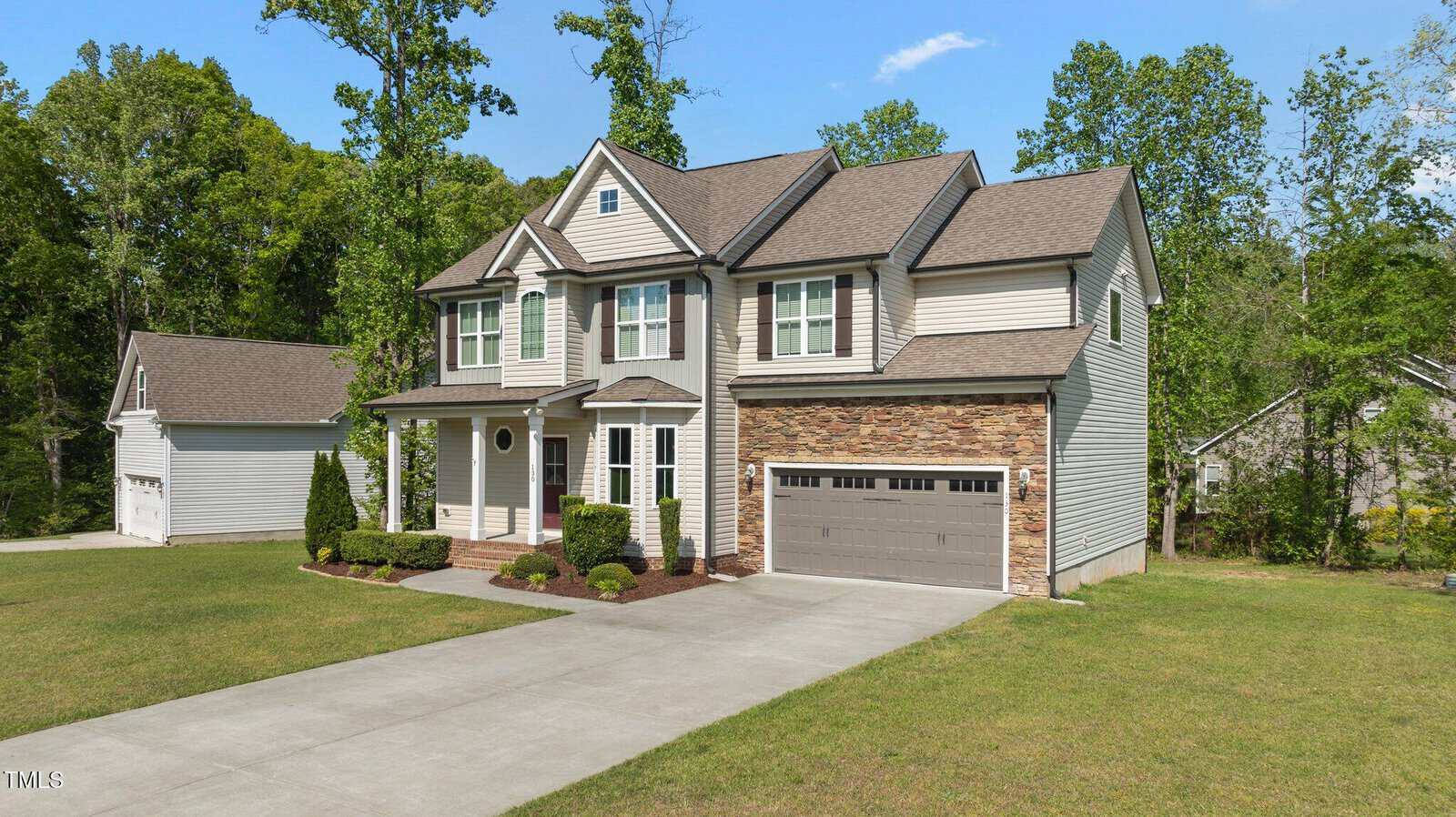 Two-story suburban house with beige siding, partial stone facade, attached two-car garage, manicured lawn, driveway, and shrubs. Surrounded by trees under a clear blue sky.