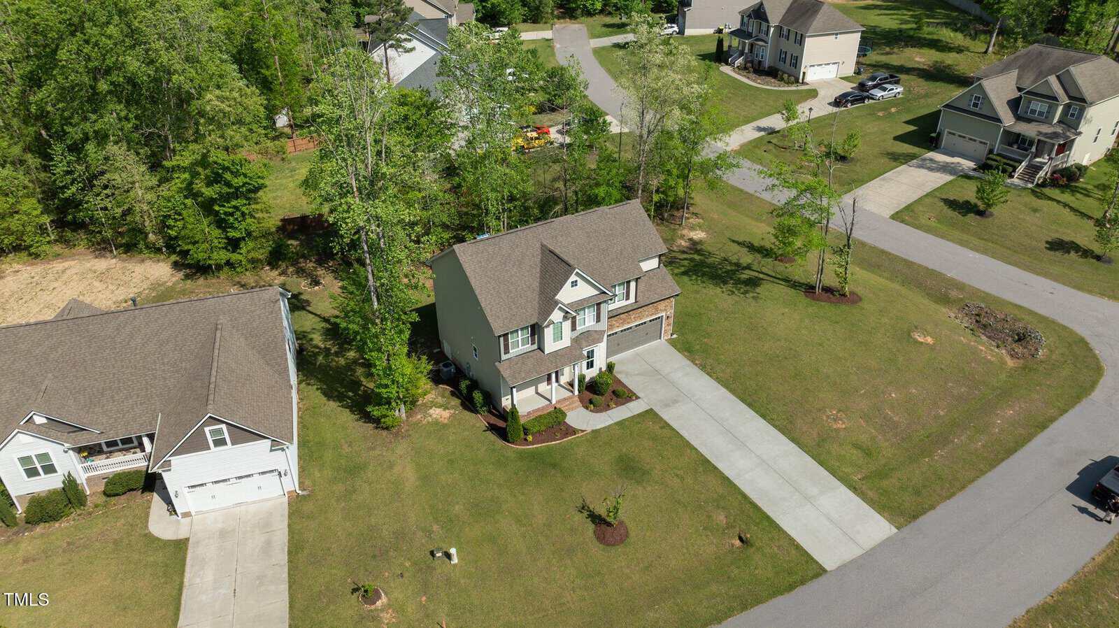 Aerial view of a two-story house with an attached garage and driveway, surrounded by a well-kept lawn and trees, located in a suburban neighborhood with nearby homes and roads.