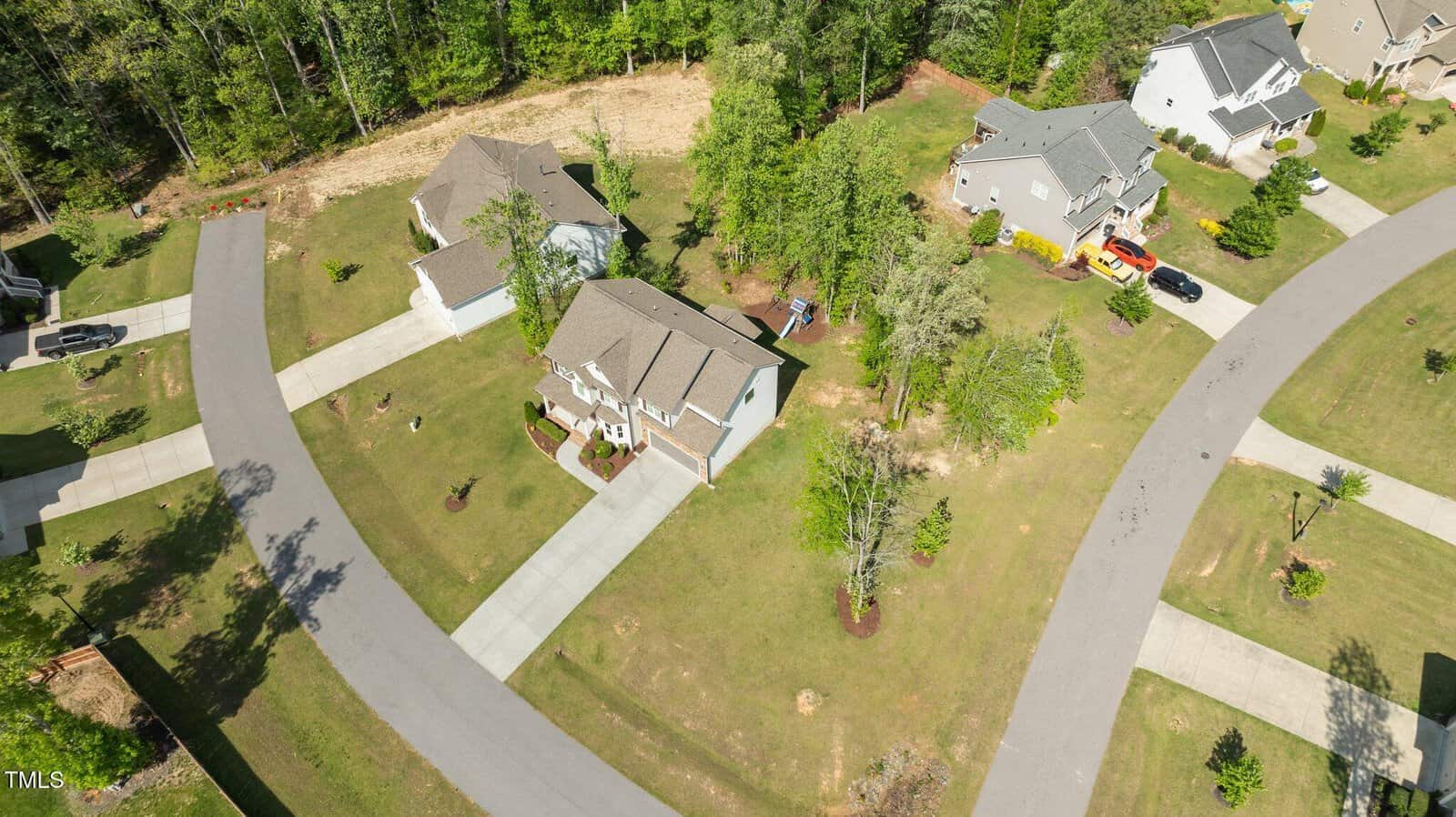 Aerial view of a suburban neighborhood showing several houses with driveways, green lawns, and surrounding trees. Curved roads pass through the area, and the properties are spaced apart with landscaped yards and sidewalks.