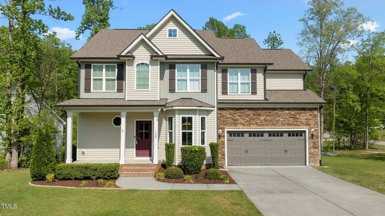 Two-story beige suburban house with stone accents, a covered front porch, dark shutters, a two-car garage, landscaped front yard, and a concrete driveway, surrounded by green trees and grass.