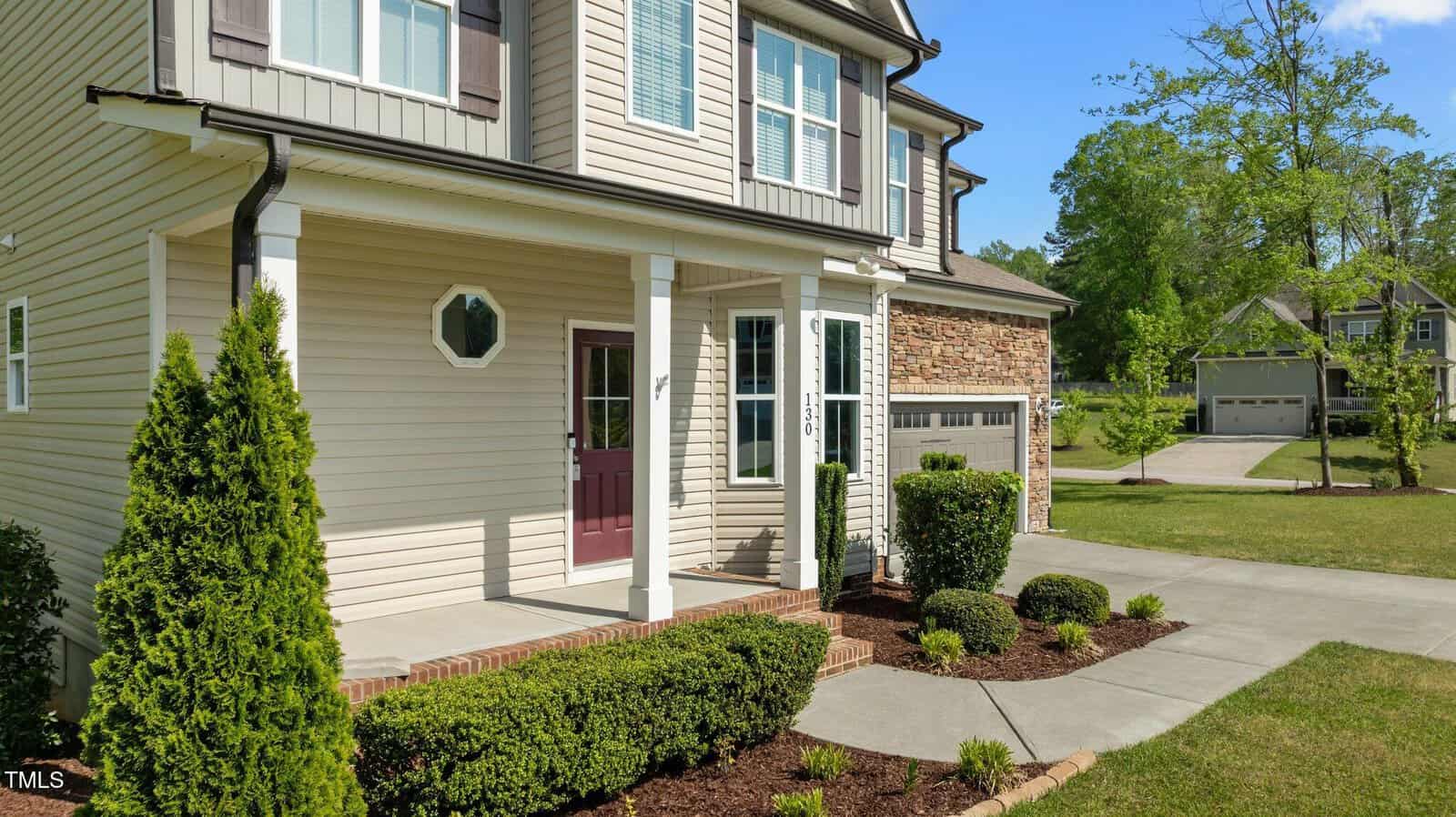 Two-story beige house with a red front door, white trim, attached garage, and a brick and stone exterior. Manicured bushes line the front porch, and a concrete pathway leads to the driveway on a sunny day.