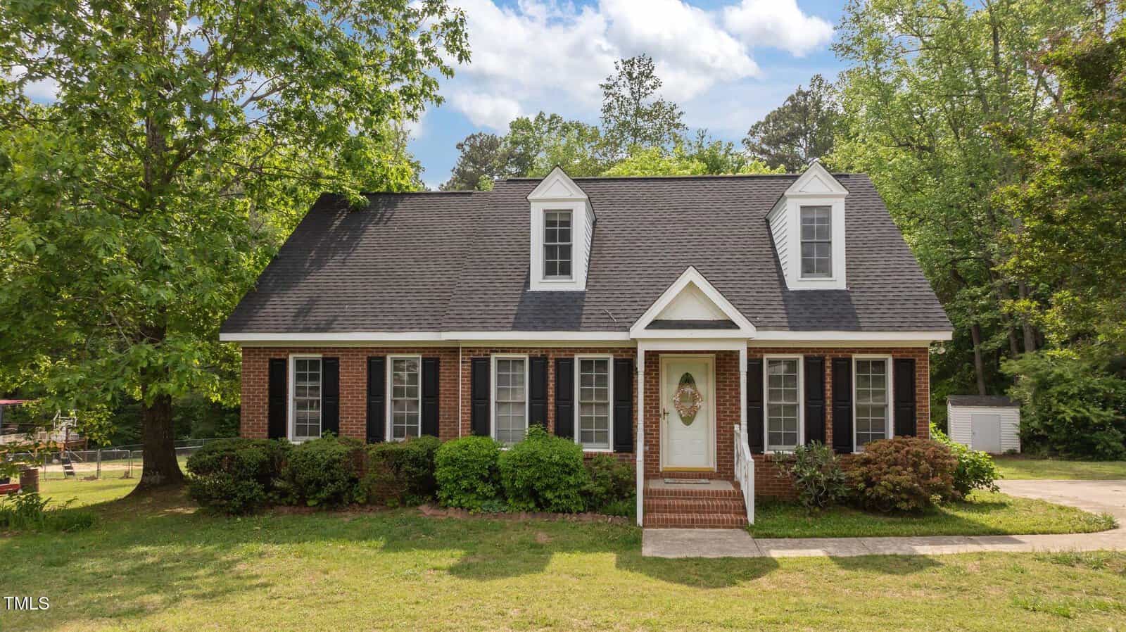 A red brick house with white trim, black shutters, and a steep dark roof featuring two dormer windows. The house has a front porch with steps, surrounded by green bushes and trees, under a partly cloudy sky.