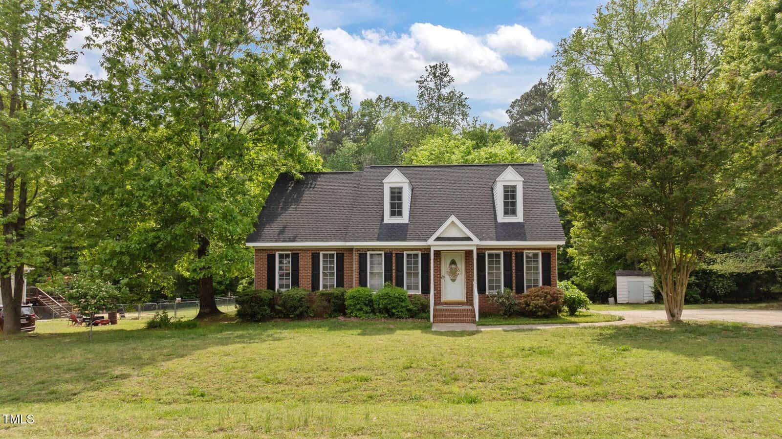 A two-story brick house with white trim, dormer windows, and a dark gray roof sits on a grassy lawn surrounded by trees under a partly cloudy sky. Steps lead up to the front door with sidelights.