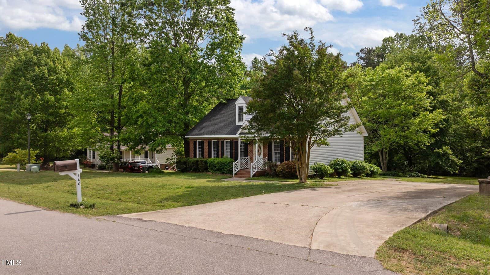 A suburban house with white siding, dark roof, and brick steps sits behind a large curved driveway. Mature trees and green lawn surround the home, with a white mailbox by the street in the foreground.