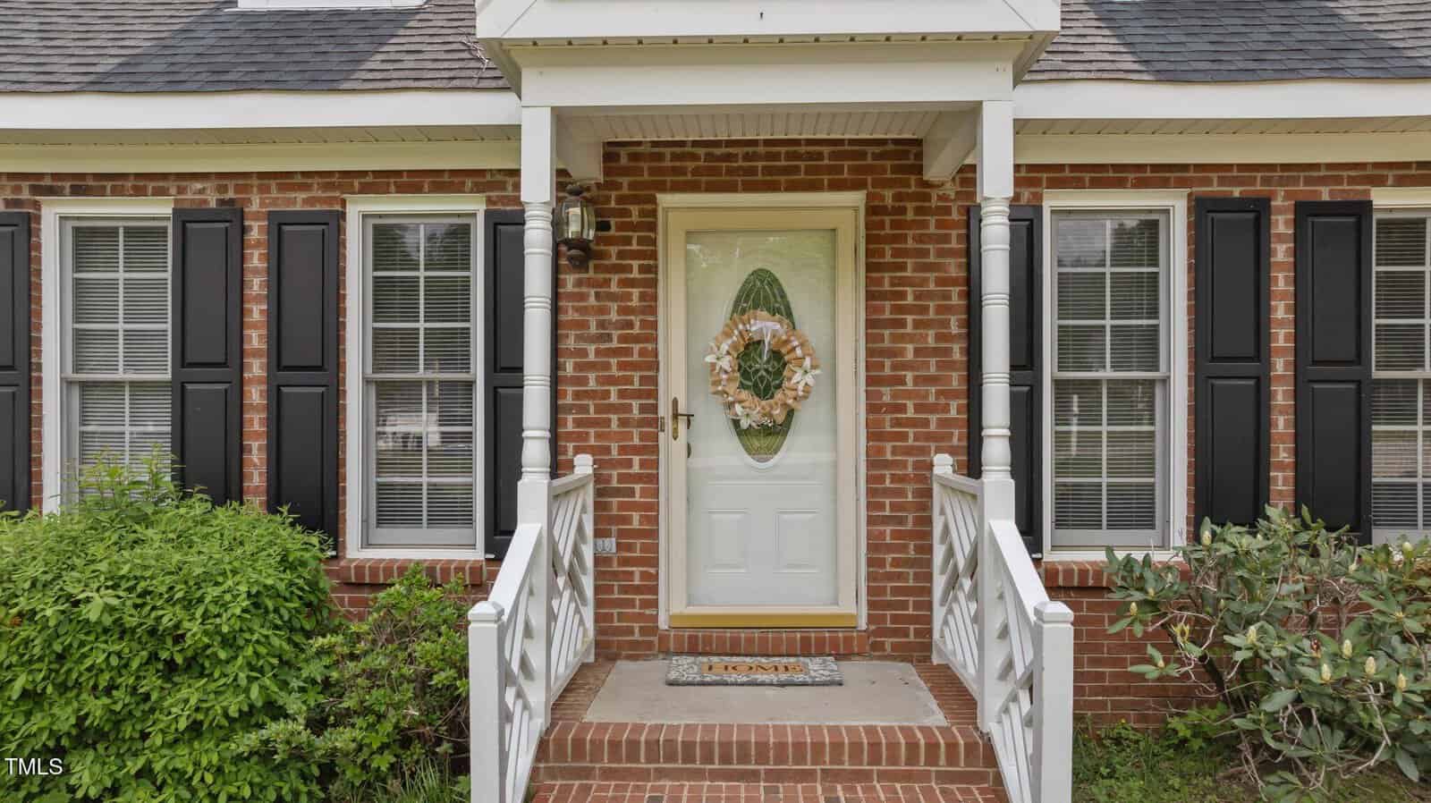 A brick house with black shutters features a white front door decorated with a floral wreath, a "Home" doormat, and a small covered porch with white railings and steps, surrounded by green bushes.