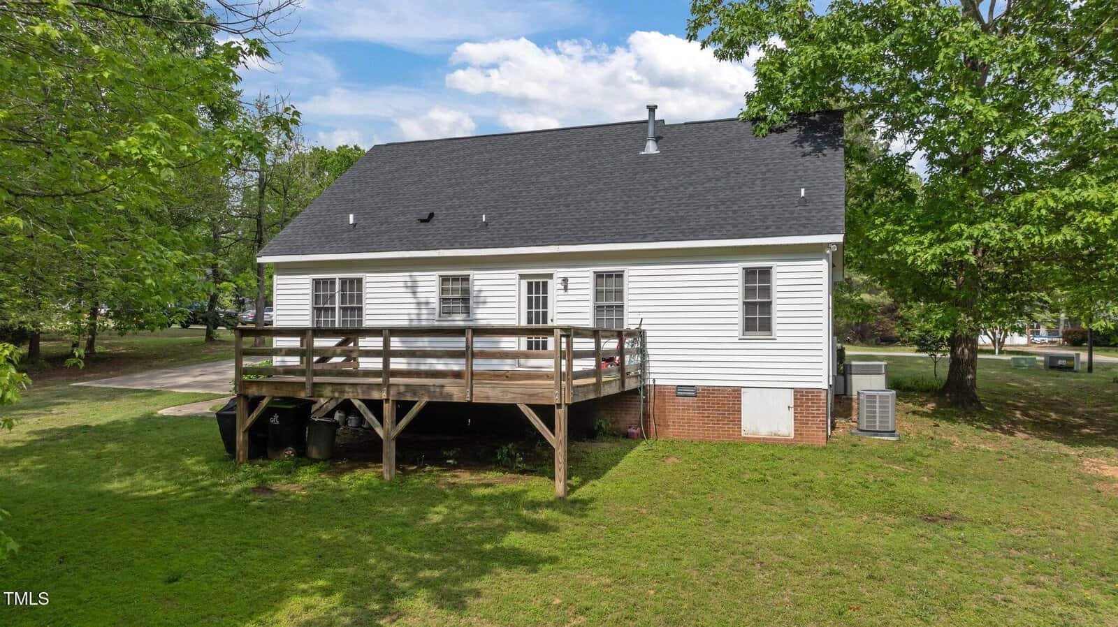 A white house with a dark shingled roof and several windows, featuring a wooden deck and stairs leading to a grassy backyard surrounded by trees under a partly cloudy sky.