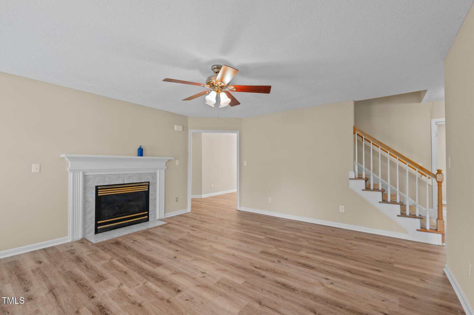 A spacious living room with light wood flooring, beige walls, a white fireplace with a black and gold trim, a ceiling fan with lights, and a staircase with a wooden handrail leading upstairs.