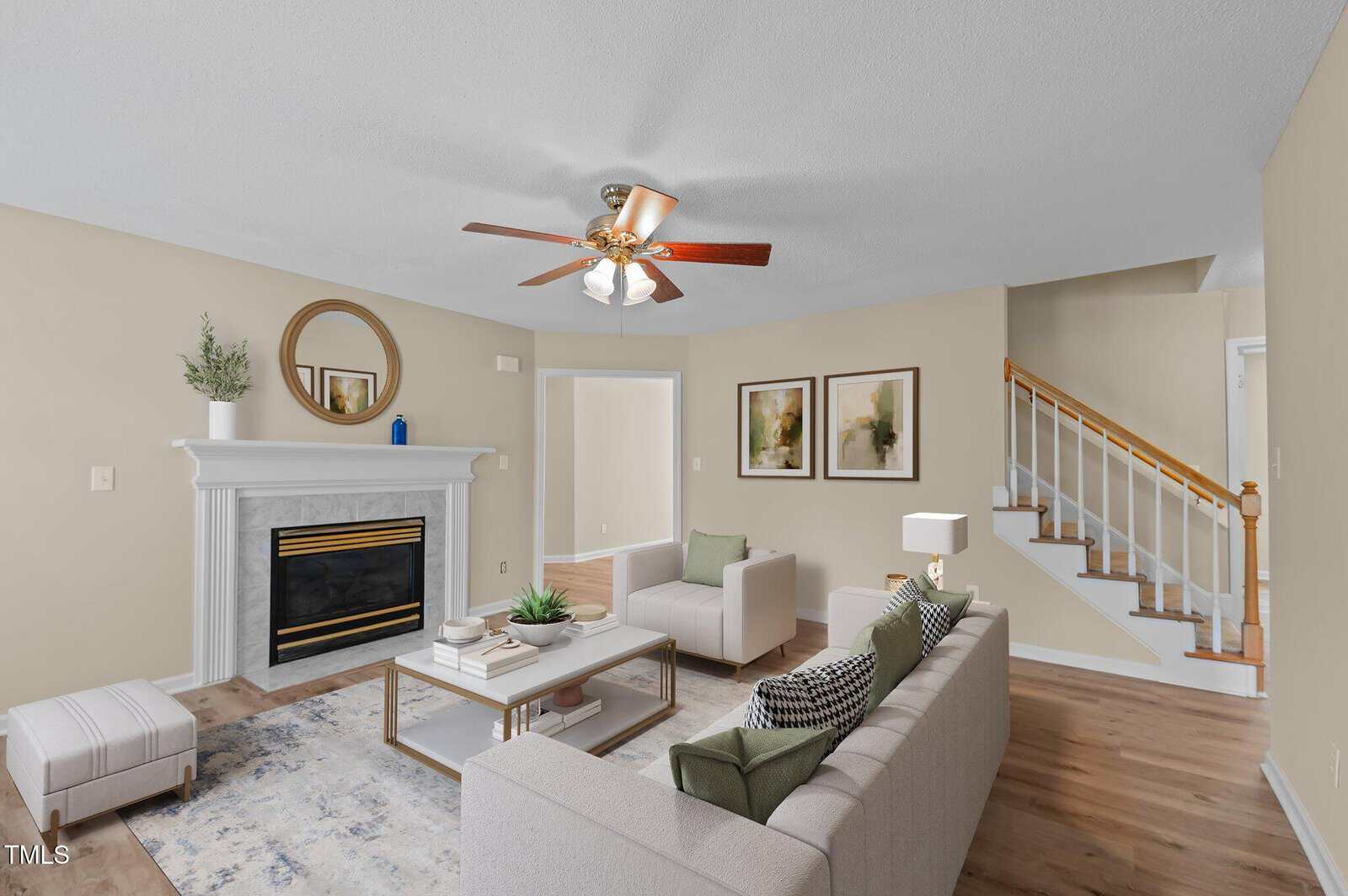 Living room with a ceiling fan, beige walls, a white fireplace, two armchairs, a sofa, coffee table, two framed prints, and a staircase with wooden steps and white railings. A small rug and pouf are on the wooden floor.