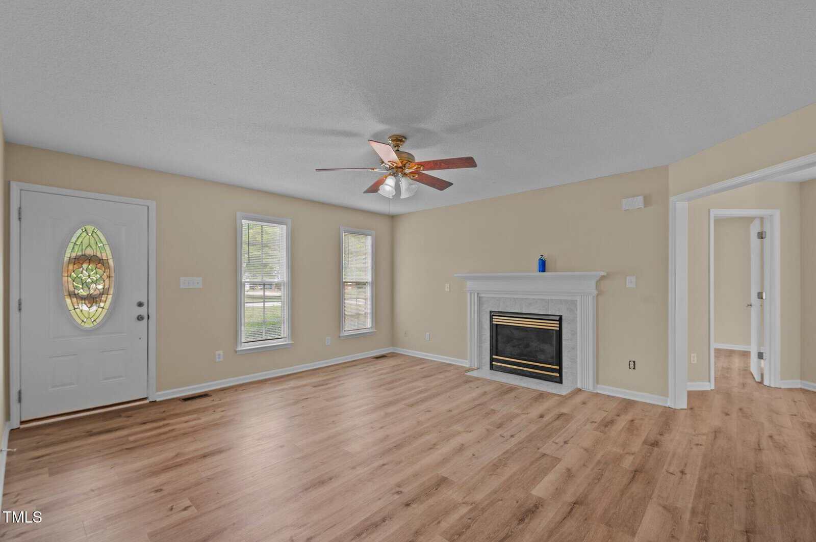 Empty living room with light wood flooring, beige walls, white trim, a ceiling fan, two front windows, a white front door with an oval glass panel, a white fireplace, and two open doorways to adjacent rooms.