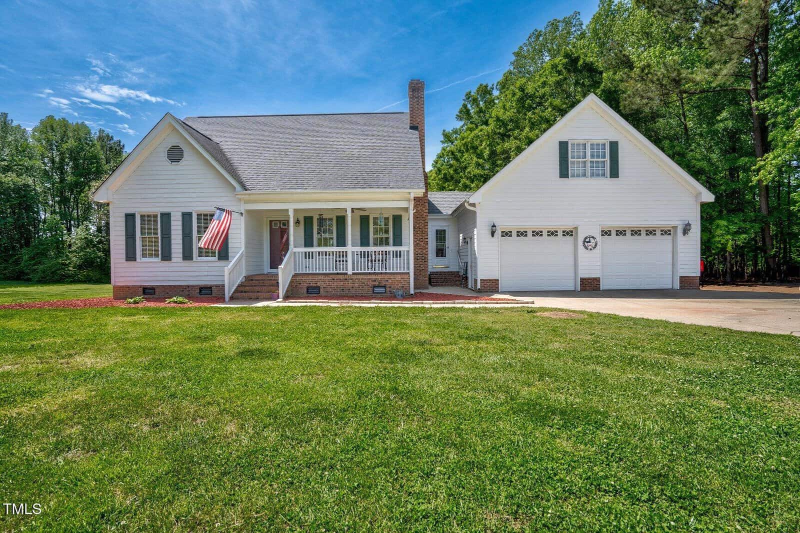 A white single-story house with a covered front porch, an American flag, and a two-car garage is located at 1576 Cheves Road in Zebulon. The home sits on a large green lawn with trees in the background under a clear blue sky.
