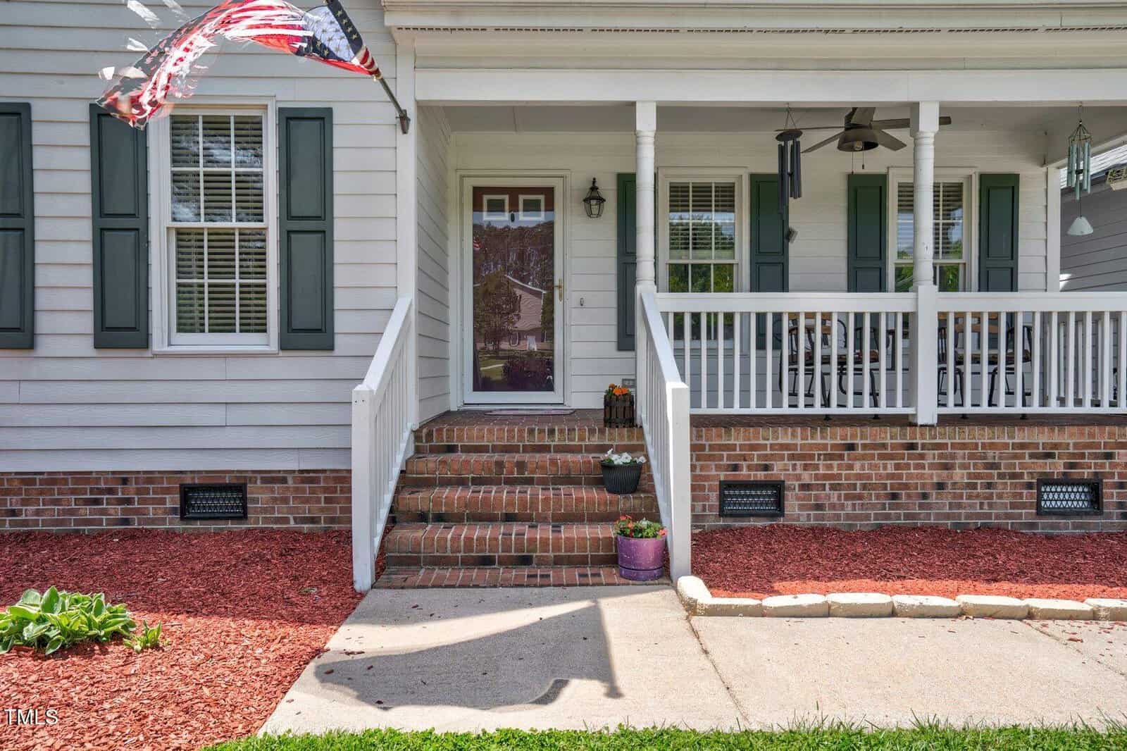A brick porch with white railings and columns leads to the front door of a house. Two flower pots are on the steps, and a flag is flying. The porch has seating, and the yard is covered with red mulch.
