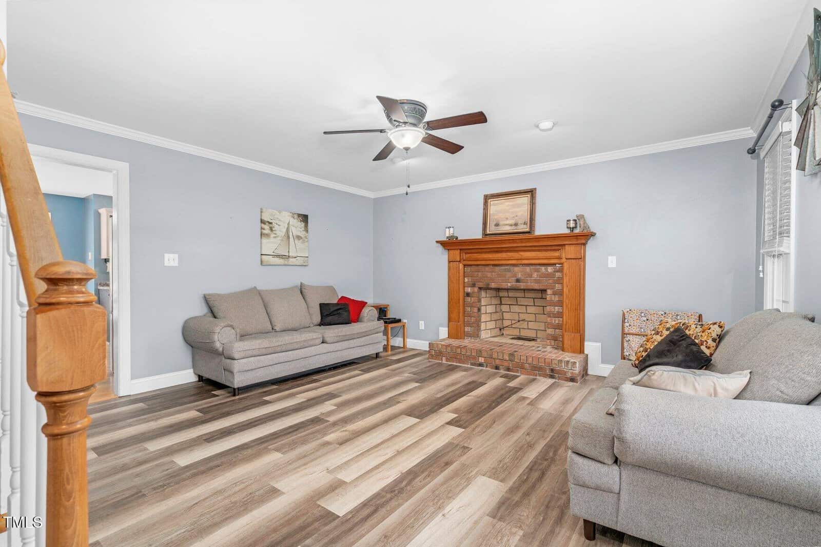Living room with wood-look flooring, gray walls, two gray sofas, a wooden fireplace with brick detail, ceiling fan with light, and a window with curtains. Artwork and a basket are beside the fireplace. Stairs are visible on the left.