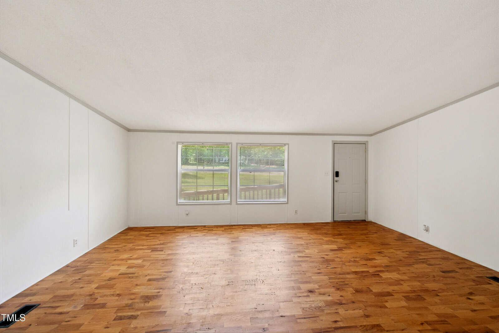 Empty room with white walls, wood flooring, two large windows, and a single door. The space is well-lit by natural light, with a view of grass and trees outside the windows.