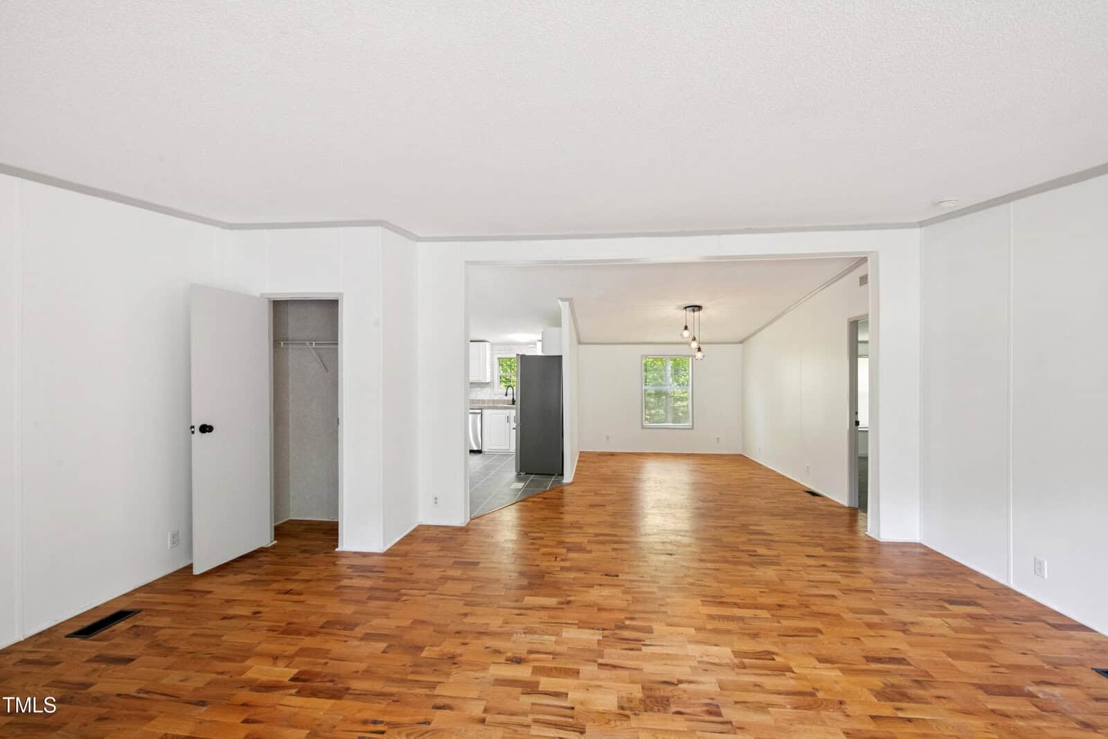 A spacious, empty room with white walls, light wood flooring, and a small open closet. The room connects to a kitchen area, visible in the background, and features a window and a hanging ceiling light.