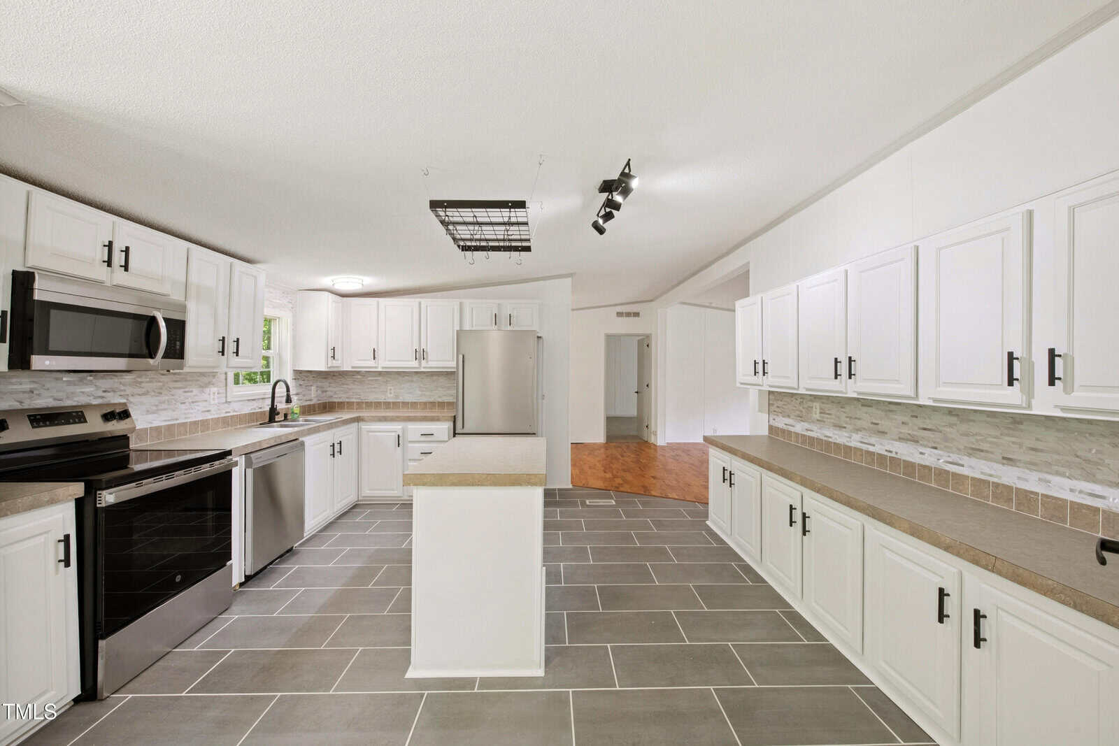 Bright kitchen with white cabinets, beige countertops, stainless steel appliances, gray tile flooring, and a light gray backsplash. There is a center island and track lighting on the ceiling. An open doorway leads to another room.