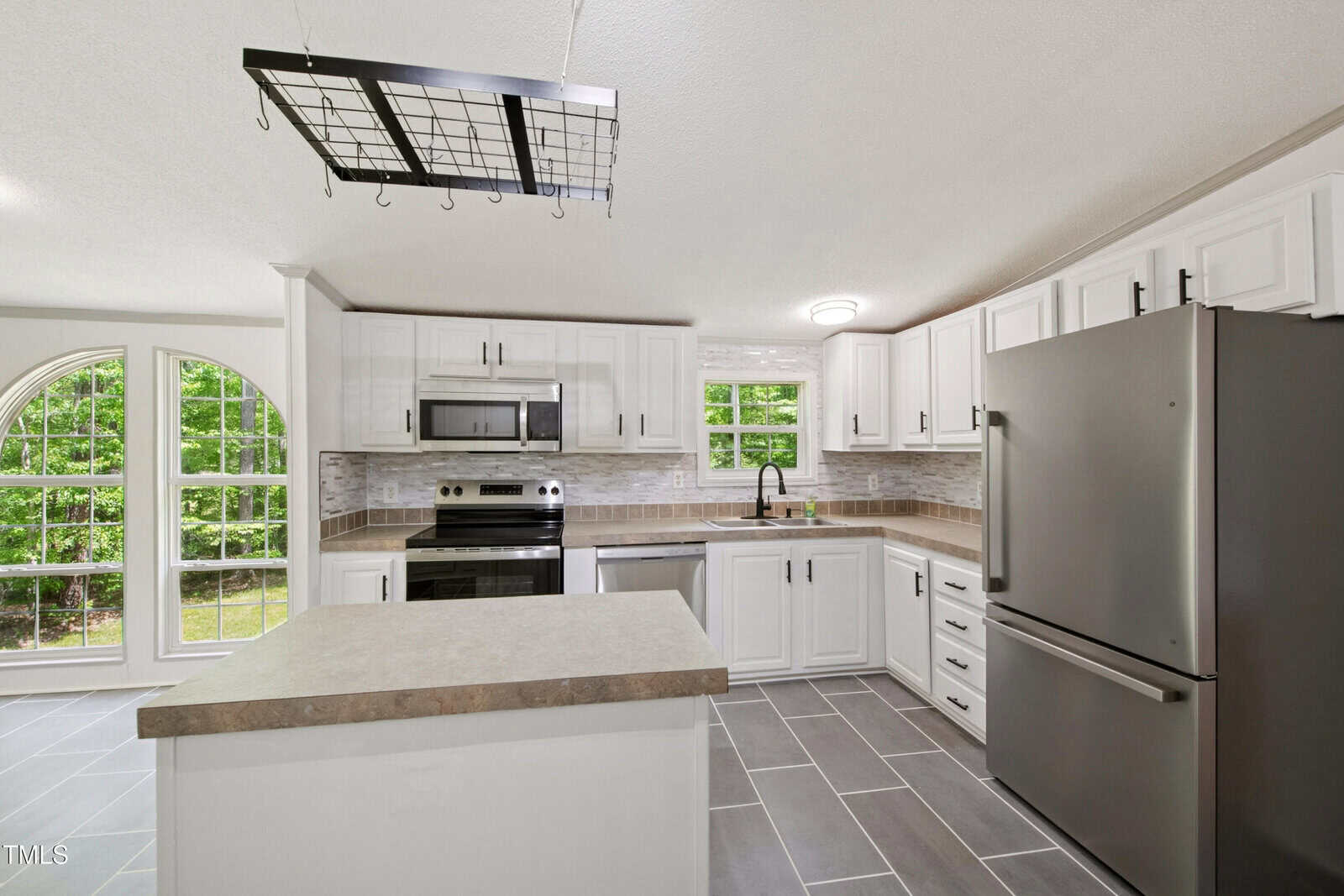 Modern kitchen with white cabinets, stainless steel appliances, a central island, gray tile flooring, tile backsplash, and a window above the sink. A pot rack hangs from the ceiling, and large windows let in natural light.