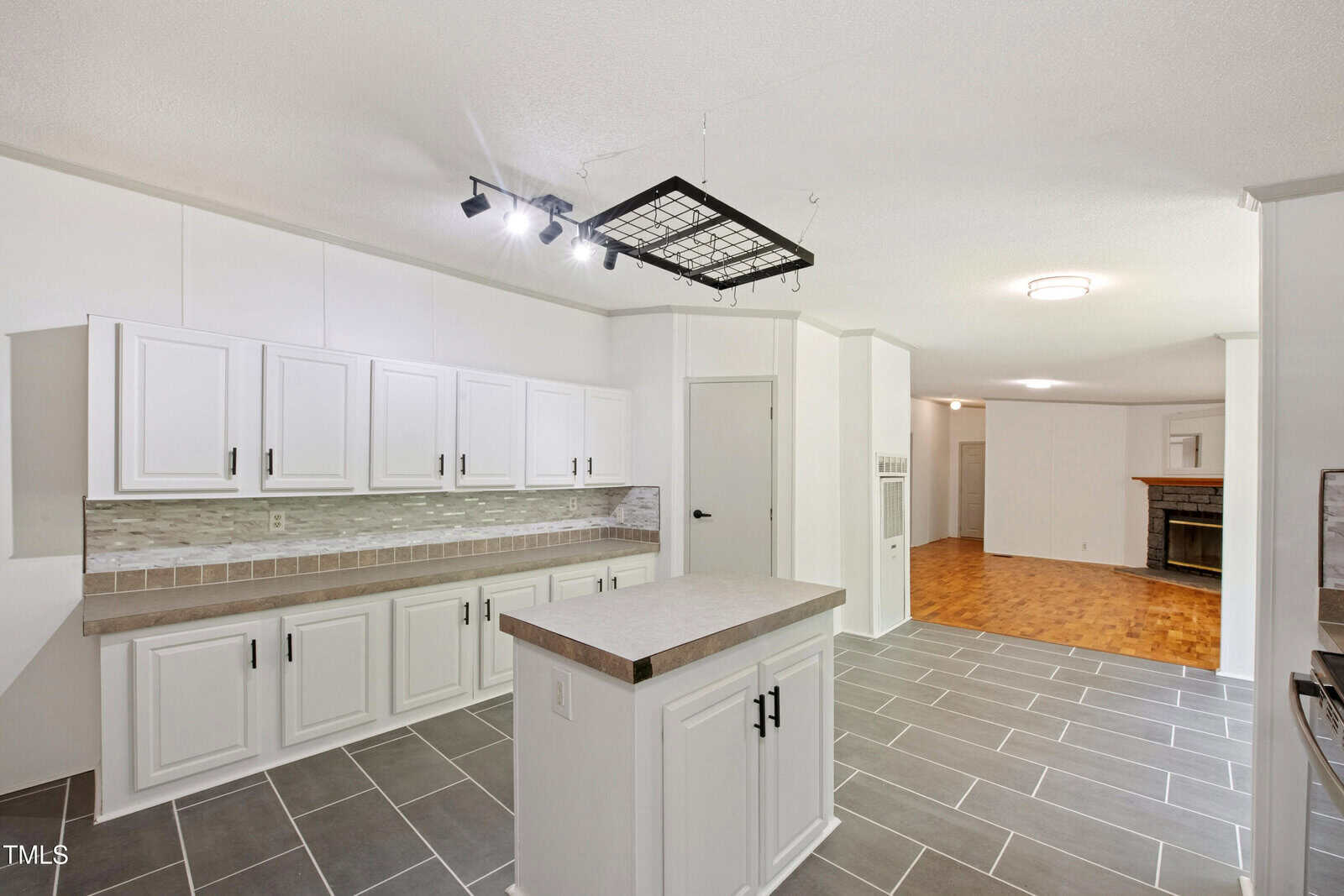 A kitchen with white cabinets, gray tile flooring, a small center island, and a tile backsplash. An open doorway leads to a living area with wood flooring and a fireplace. Black track lighting hangs above the island.