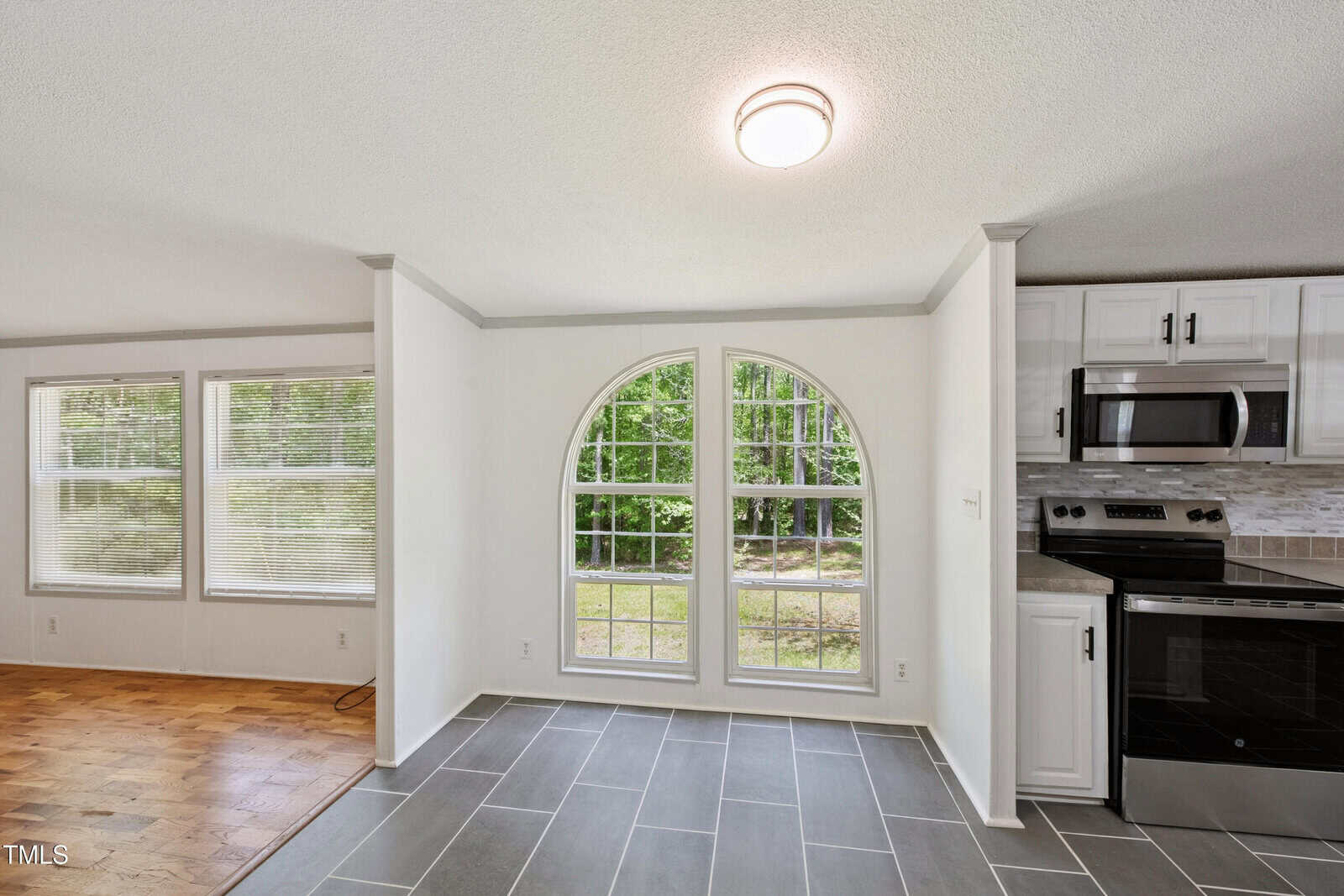 A bright room with large arched windows, slate gray tile flooring, white walls, and a view of trees outside. Part of a kitchen with stainless steel appliances and white cabinets is visible to the right.