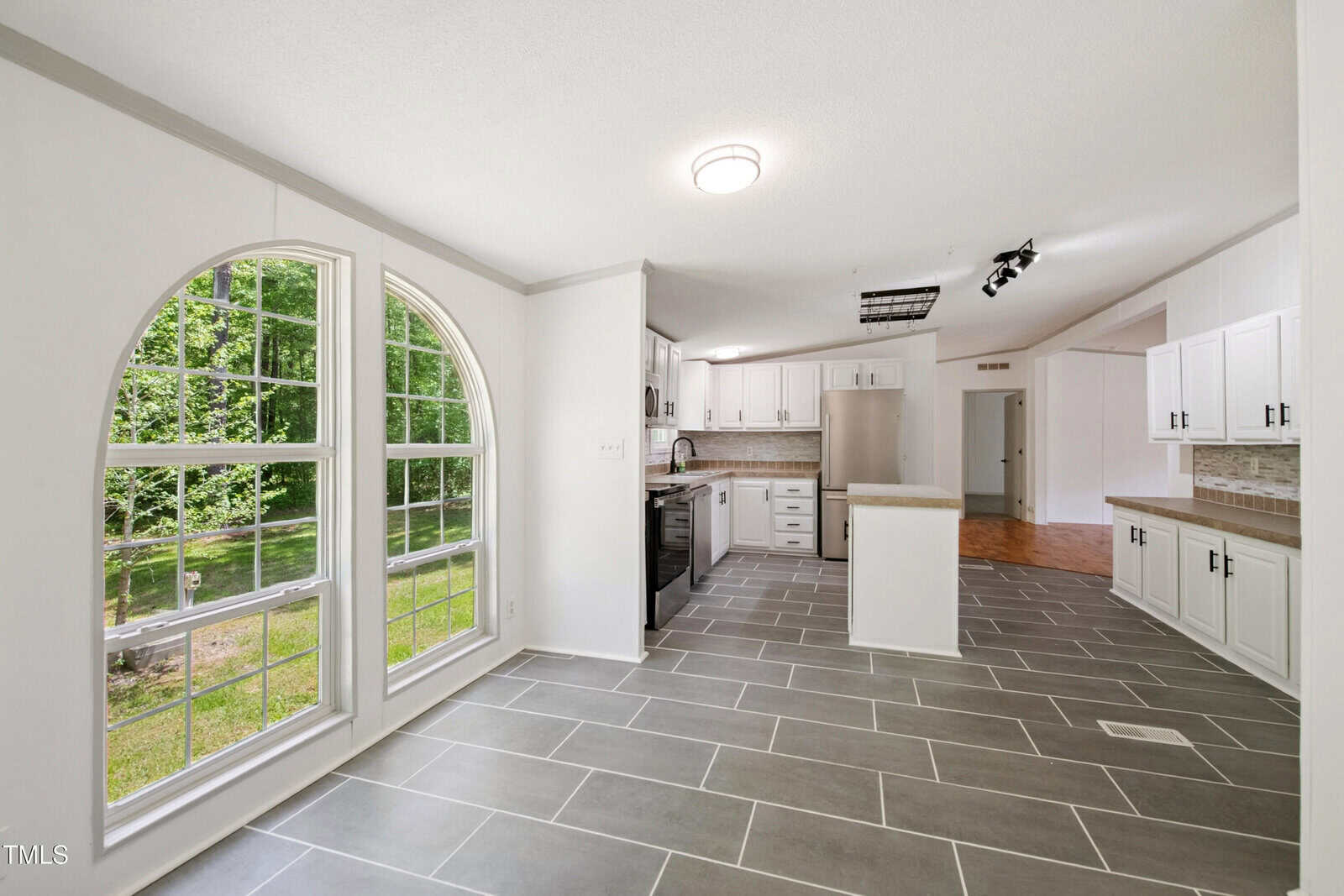 Bright kitchen with white cabinets, stainless steel appliances, gray tile floor, and a large arched window overlooking a green yard. The room is spacious and connects to other areas of the home.