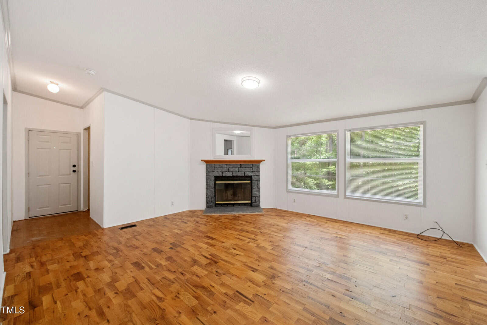 A spacious living room with light-colored walls, hardwood flooring, a wood-framed fireplace, three large windows with blinds, and a closed front door visible to the left.