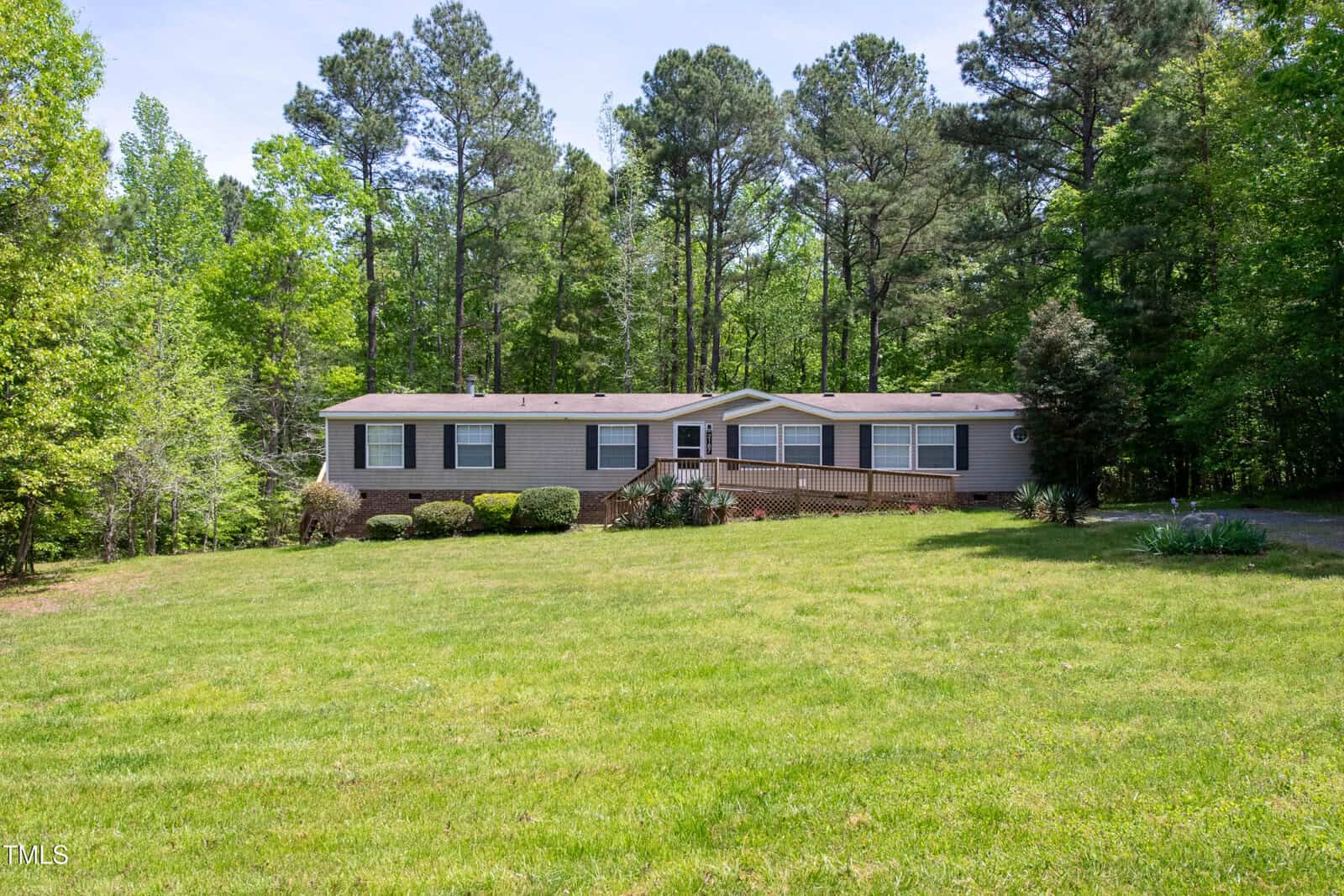 A single-story, light-colored manufactured home at 2187 Lauren Mill Drive, Oxford, features a front porch and ramp on a large, grassy lawn surrounded by tall trees and lush greenery under a clear sky.