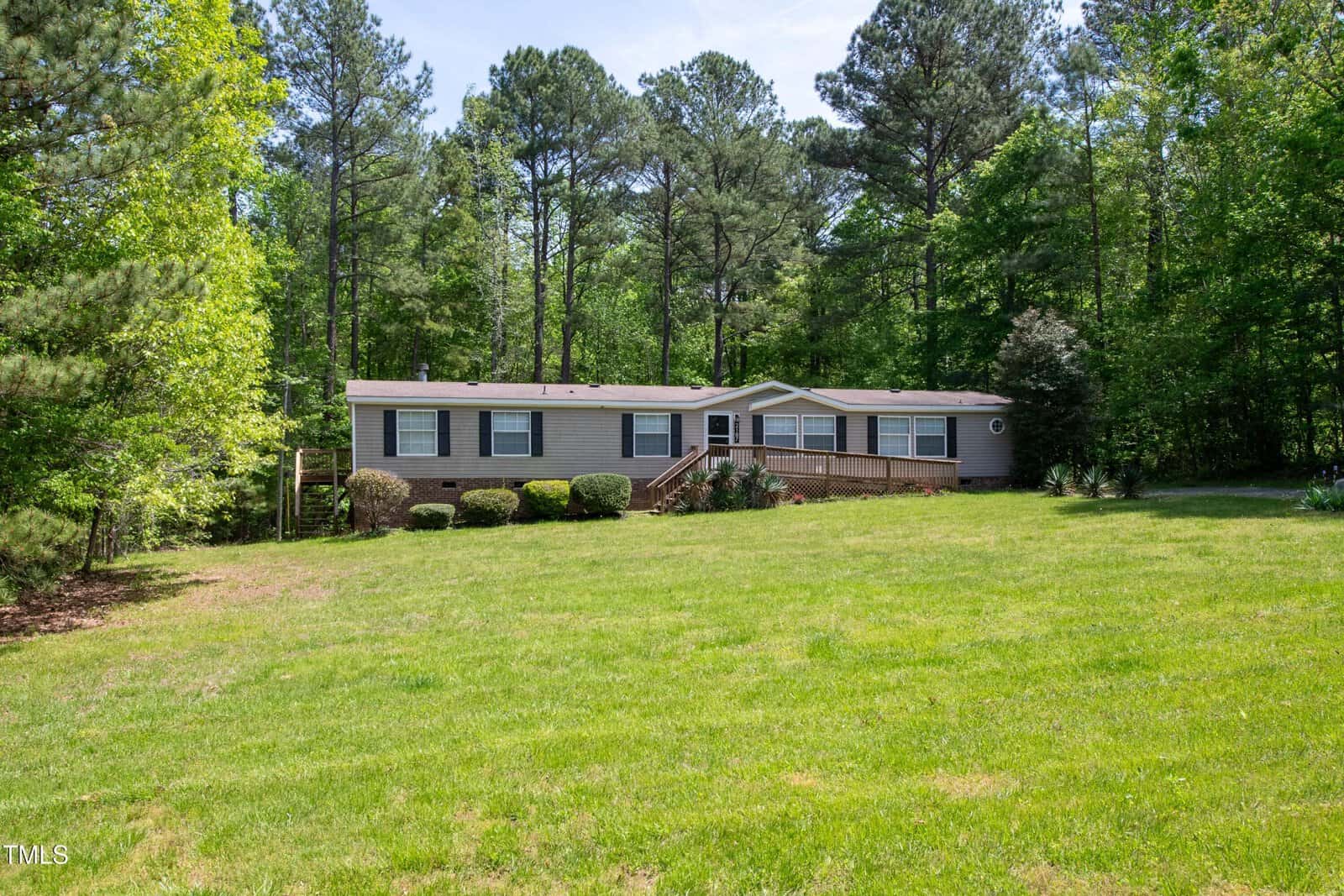 A single-story manufactured home with beige siding and brick foundation sits in a large grassy yard, surrounded by trees and shrubs, under a clear sky.