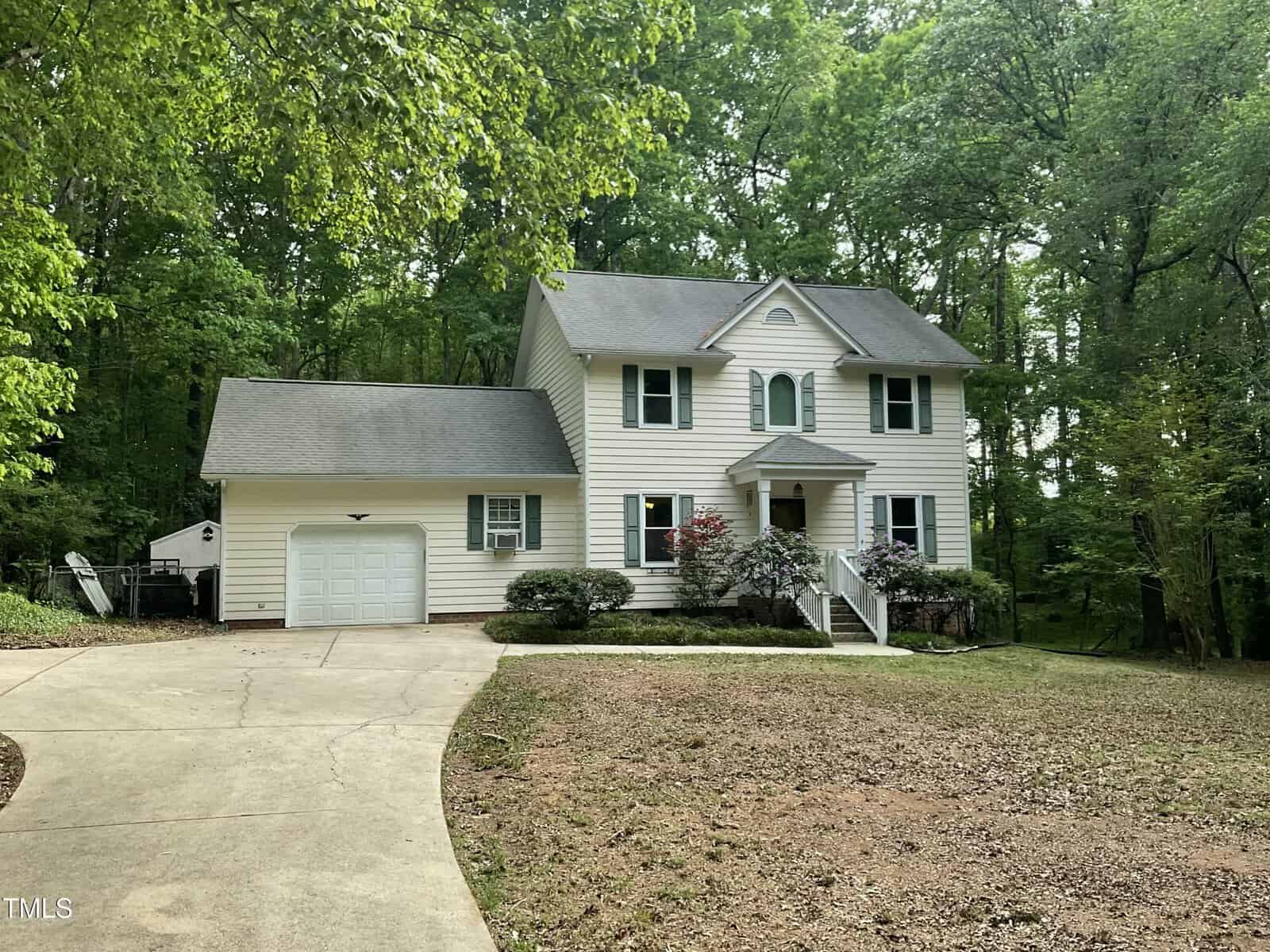 Charming two-story white house at 3648 Sleepy Hollow in Wake Forest, featuring green shutters, a one-car garage, curved driveway, small front porch with steps, and lush trees and bushes near the entrance.