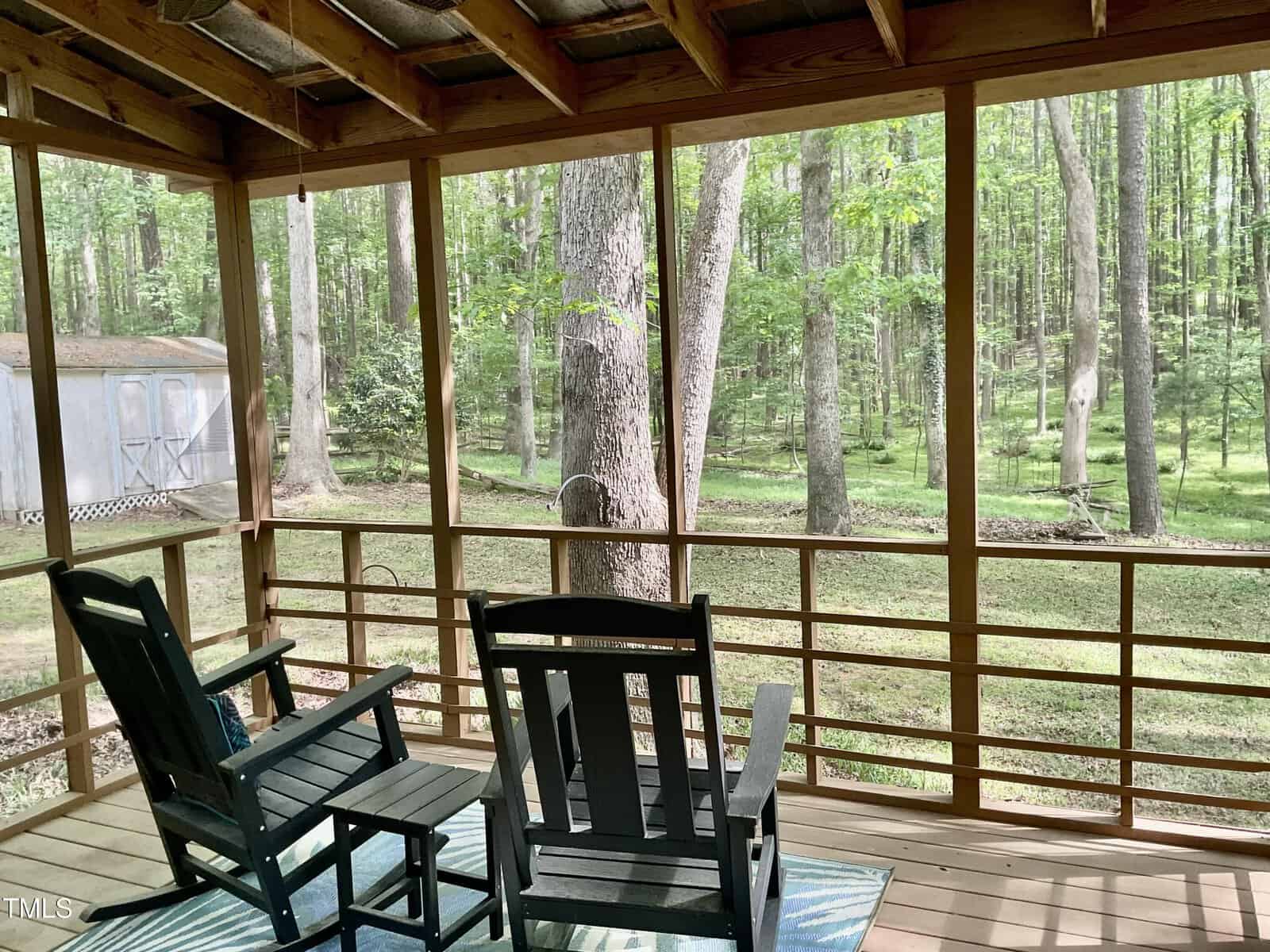 Two black rocking chairs sit on a screened-in porch overlooking a wooded backyard with tall trees and a shed in the distance. Sunlight filters through the trees, and a rug is placed under the chairs.