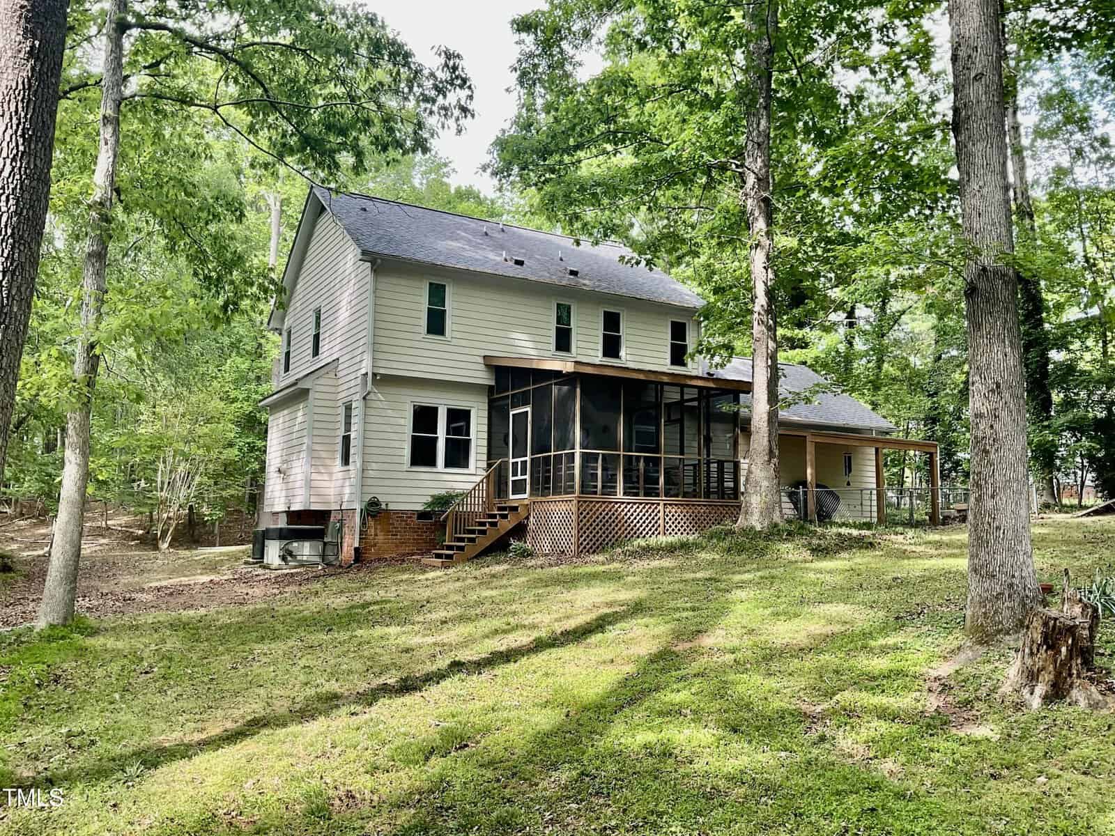 A two-story beige house with a dark gray roof sits among tall trees. The house features a screened-in porch with steps leading to a grassy yard, and a covered area on the right side. The setting is wooded and peaceful.