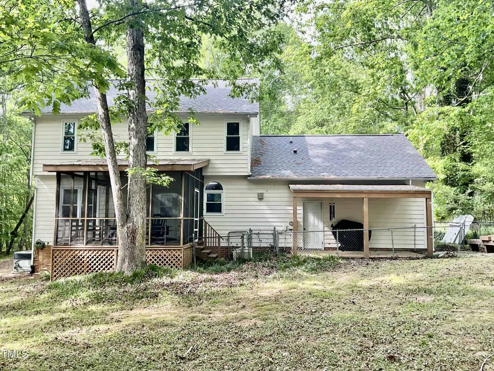 A two-story light-colored house with a screened porch, a covered carport area, fenced yard, and surrounding trees. The ground is patchy with grass and leaves, and the area appears shaded by foliage.