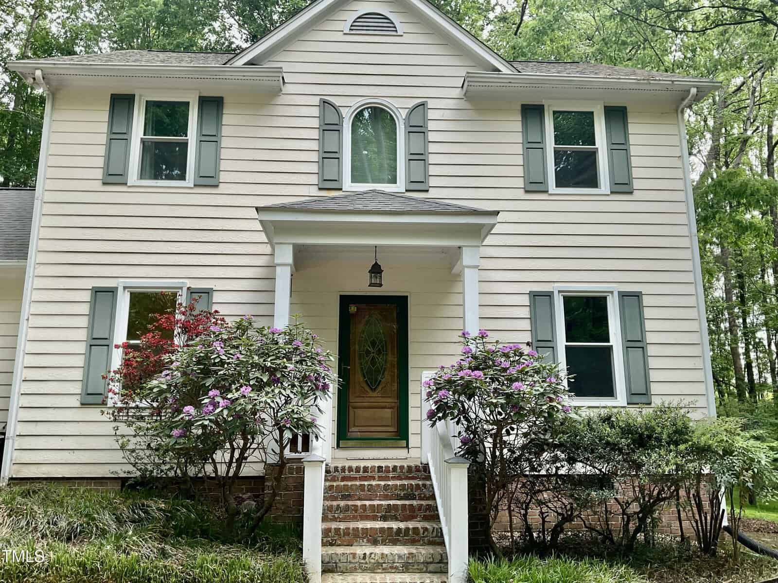 A two-story beige house with light green shutters, a wooden front door with an oval glass window, brick steps, and flowering bushes in front, surrounded by trees.