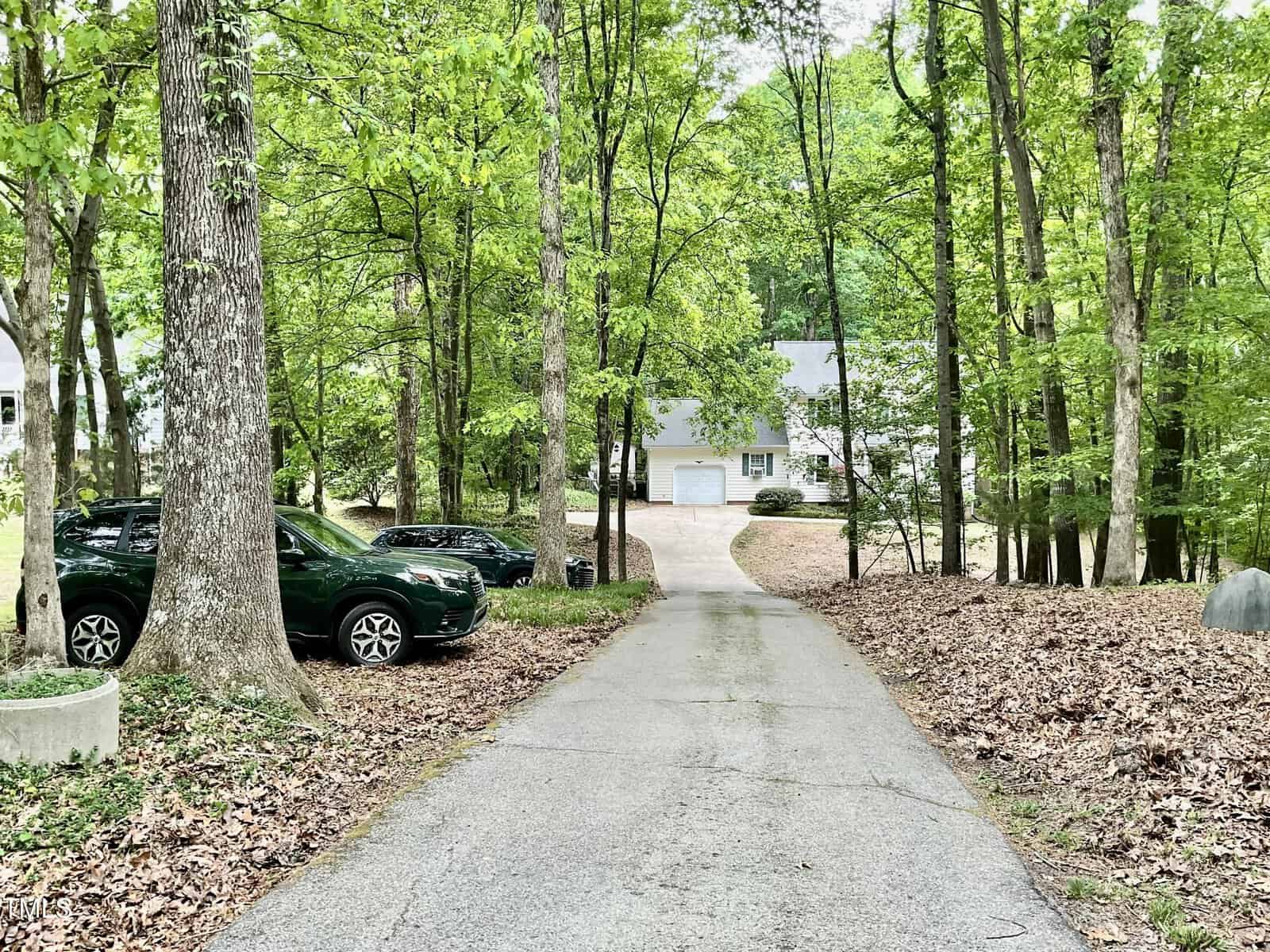 A paved driveway leads through a wooded area with green trees and parked cars on the left, ending at a white house with a two-car garage in the background. Fallen leaves cover the ground on both sides of the driveway.