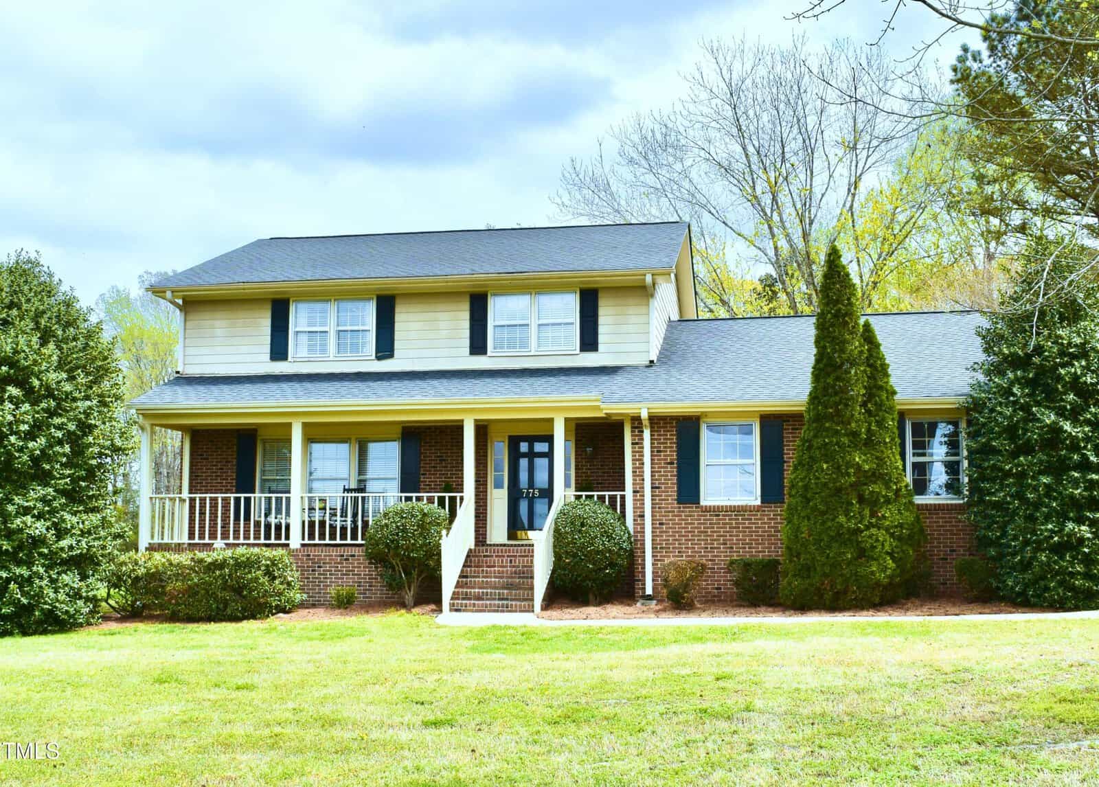 A two-story suburban house with brick and light-colored siding, black shutters, and a covered front porch. The lawn is green, with shrubs and trees surrounding the home on a partly cloudy day.