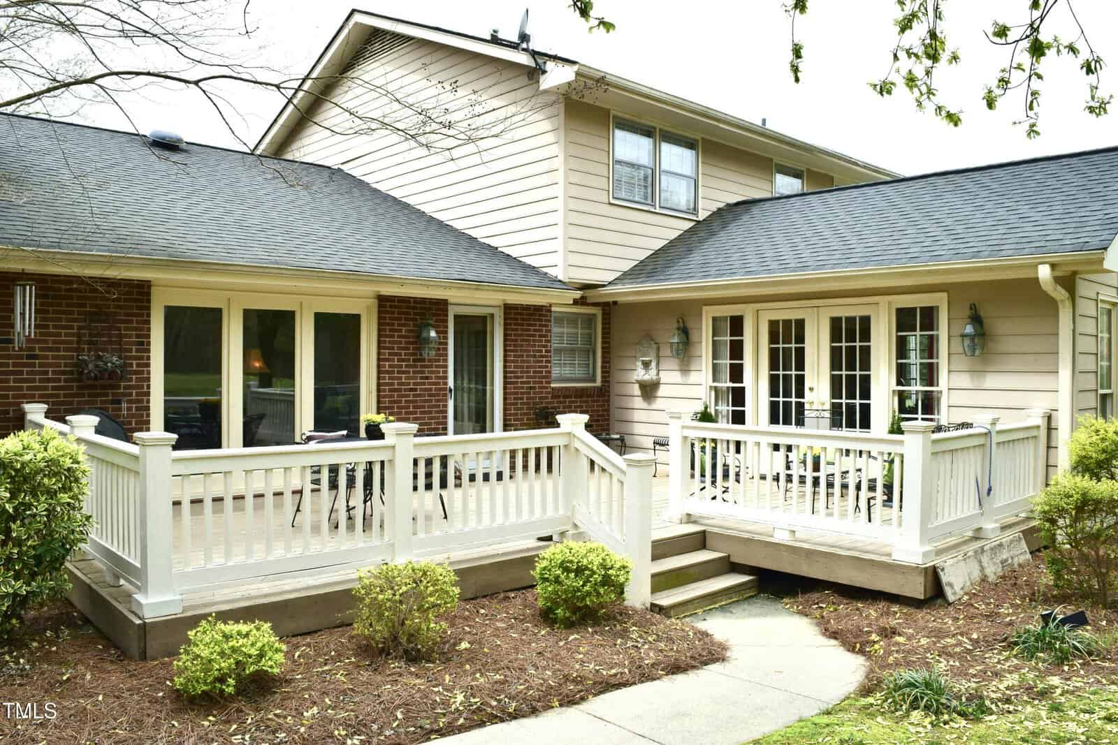 A beige two-story house with a brick accent wall features a wooden deck and white railings, accessed by steps. French doors and large windows open to the deck, surrounded by small shrubs and a walkway.