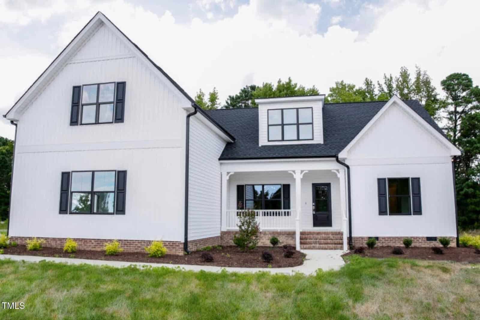 A modern white house with black shutters, a brick foundation, and a covered front porch. The yard is landscaped with small shrubs and a green lawn, with trees in the background.