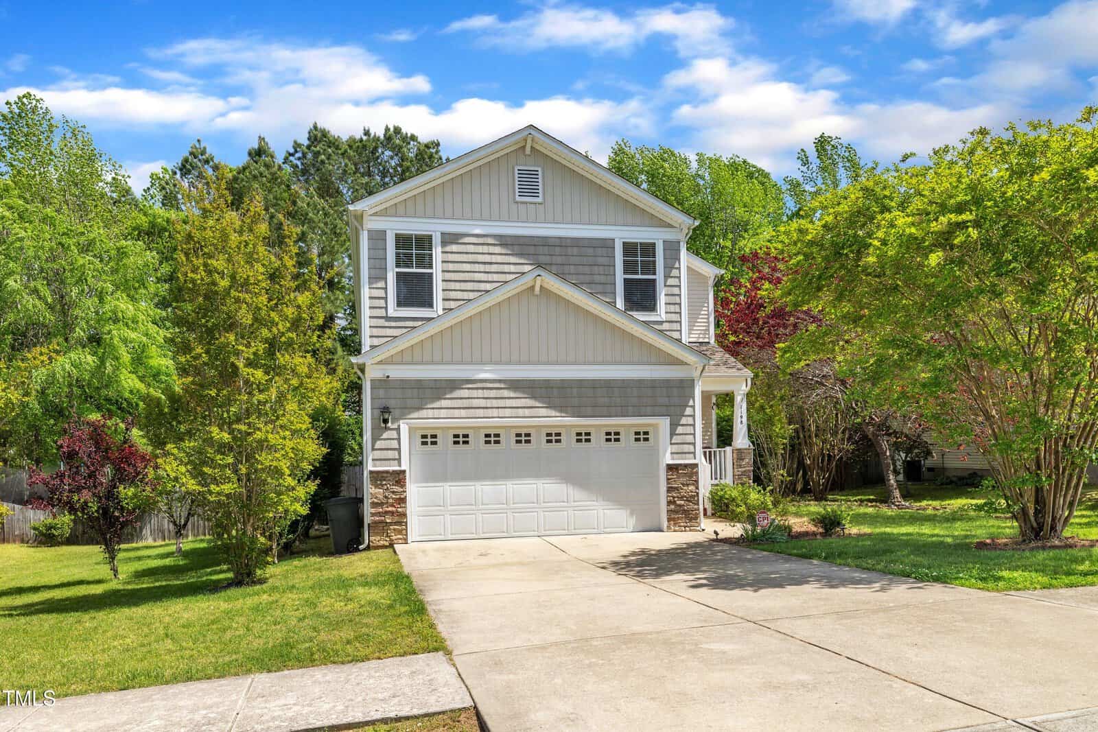 A two-story suburban house with gray siding, white trim, and an attached two-car garage sits at the end of a concrete driveway, surrounded by green trees and a well-kept lawn under a partly cloudy sky.